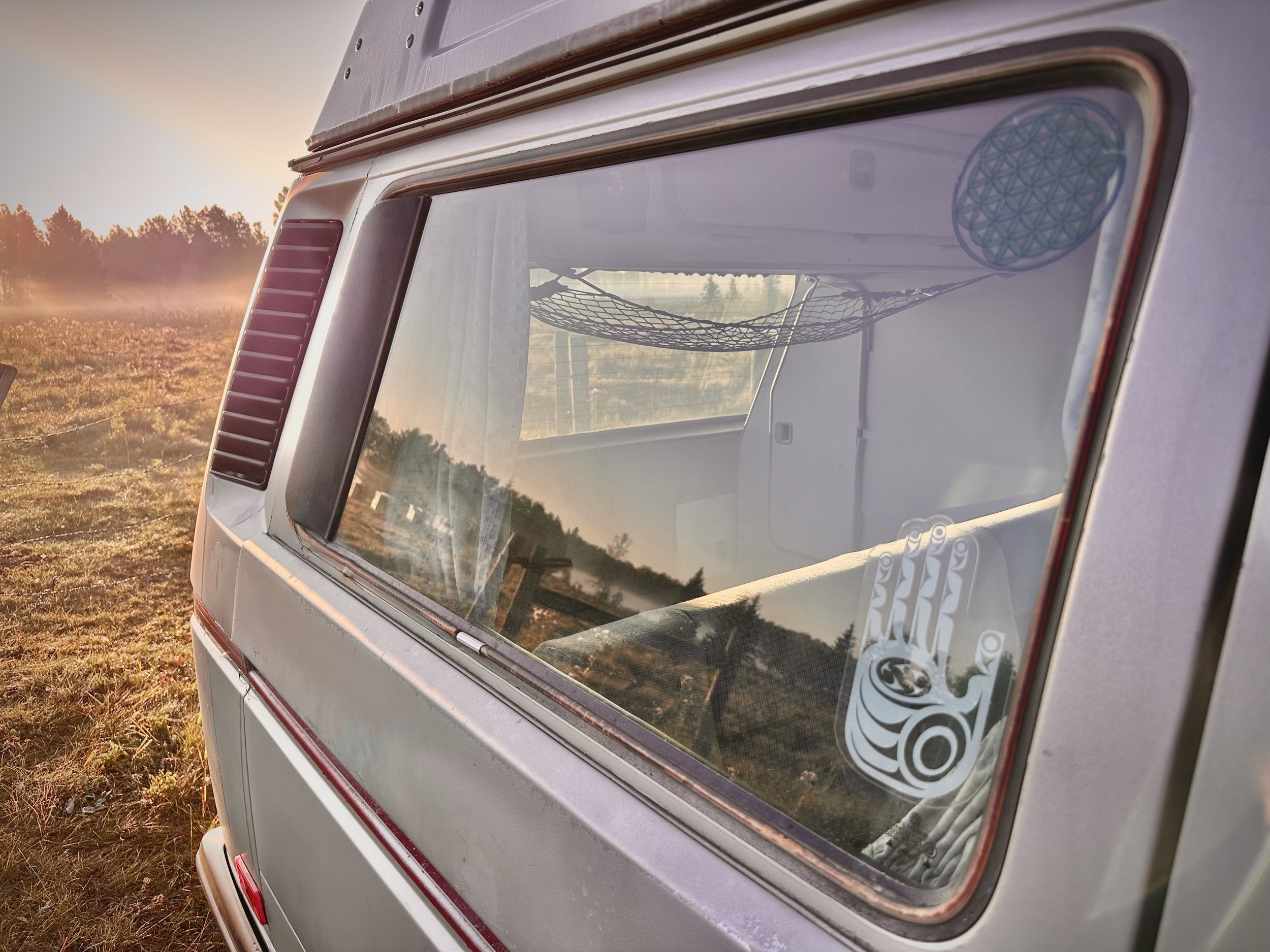 Side view of a vintage Vanagon camper van parked in an open grassy field during sunrise, with reflections of the landscape on the van's rear window.