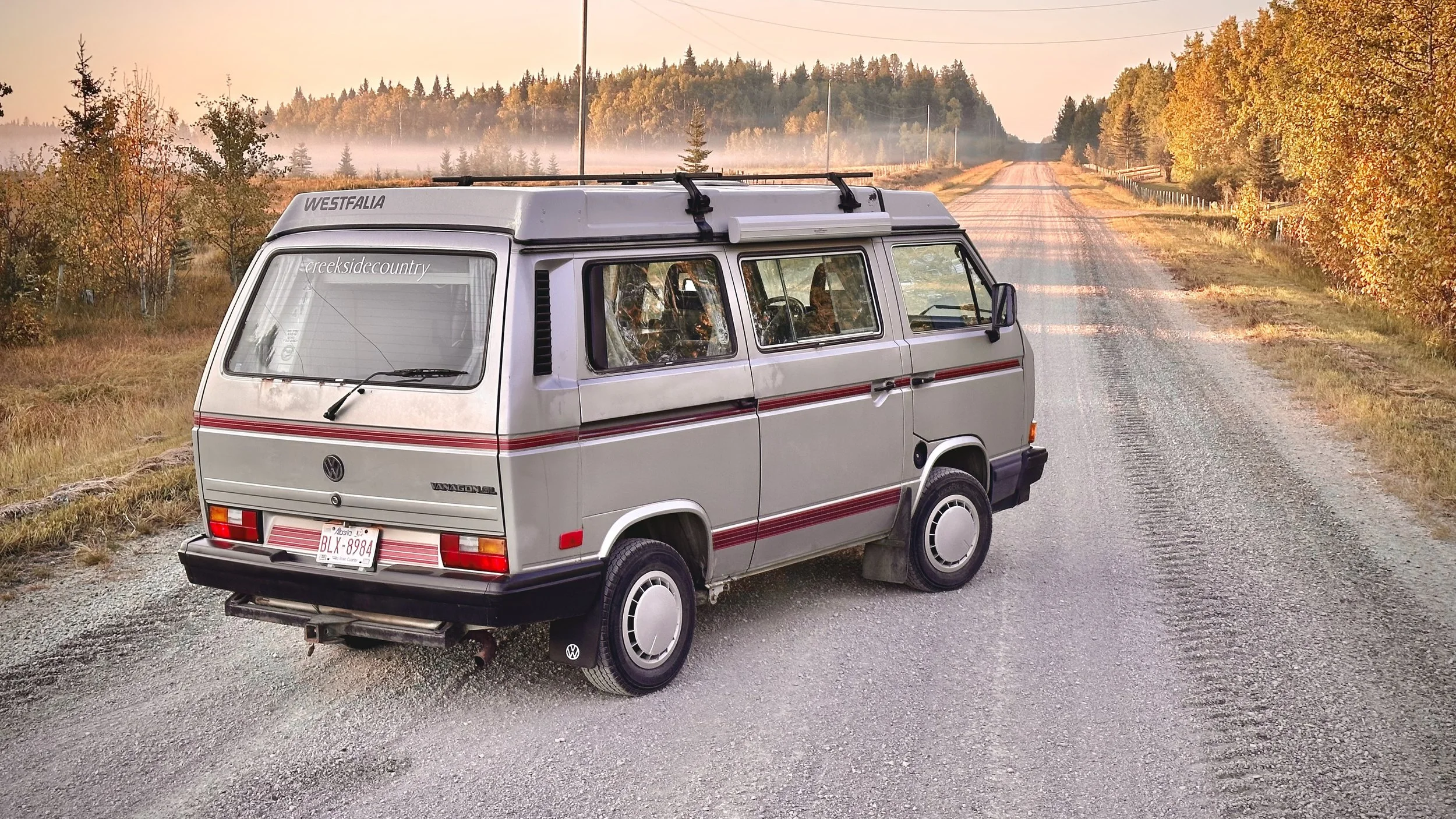 A vintage silver Volkswagen Vanagon parked on a dirt road surrounded by autumn trees, with a sunset in the background.