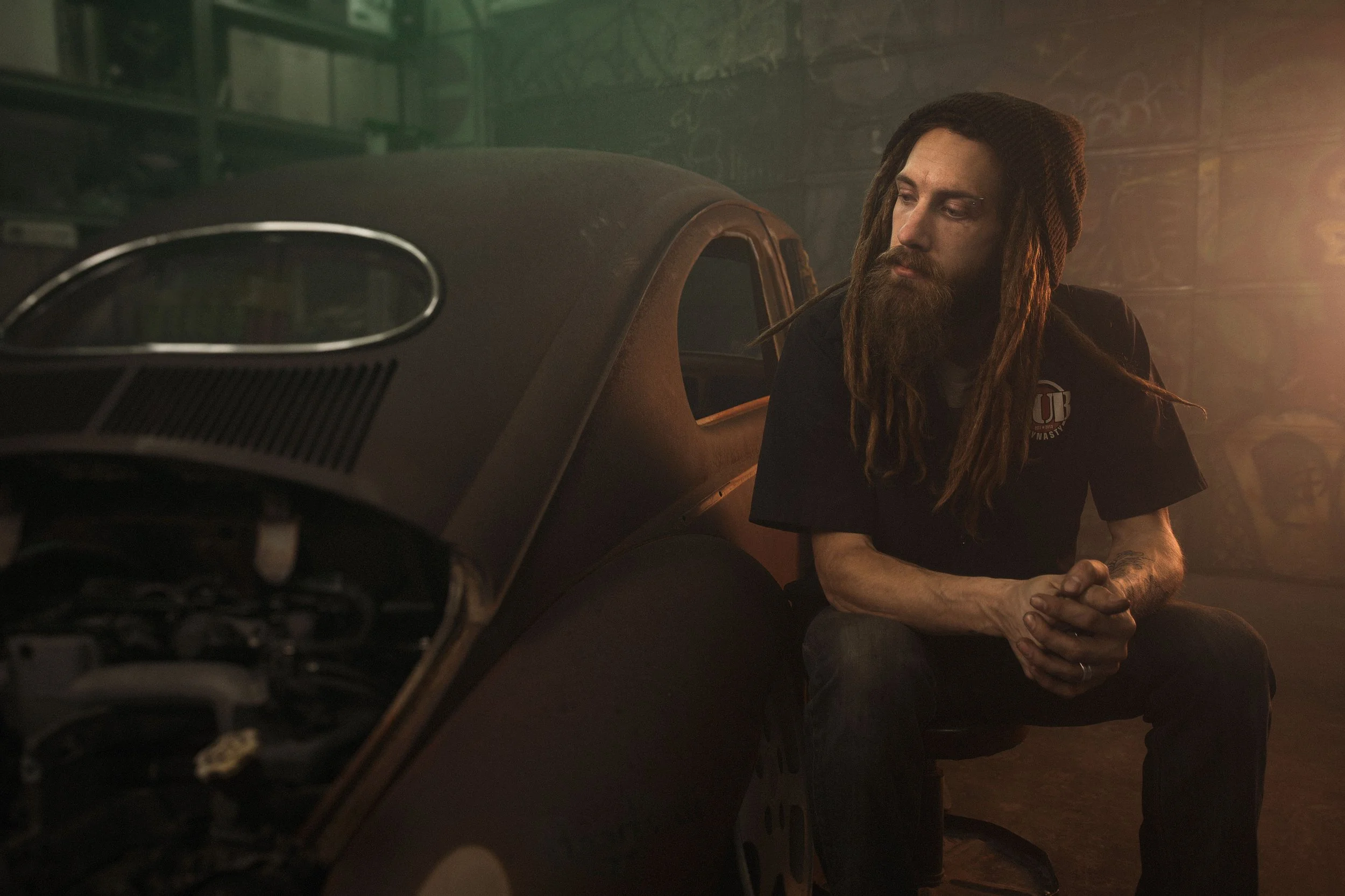 A man with long dreadlocks and a beard sits next to a vintage car in a dimly lit garage, with graffiti on the walls, wearing a black T-shirt with a logo.