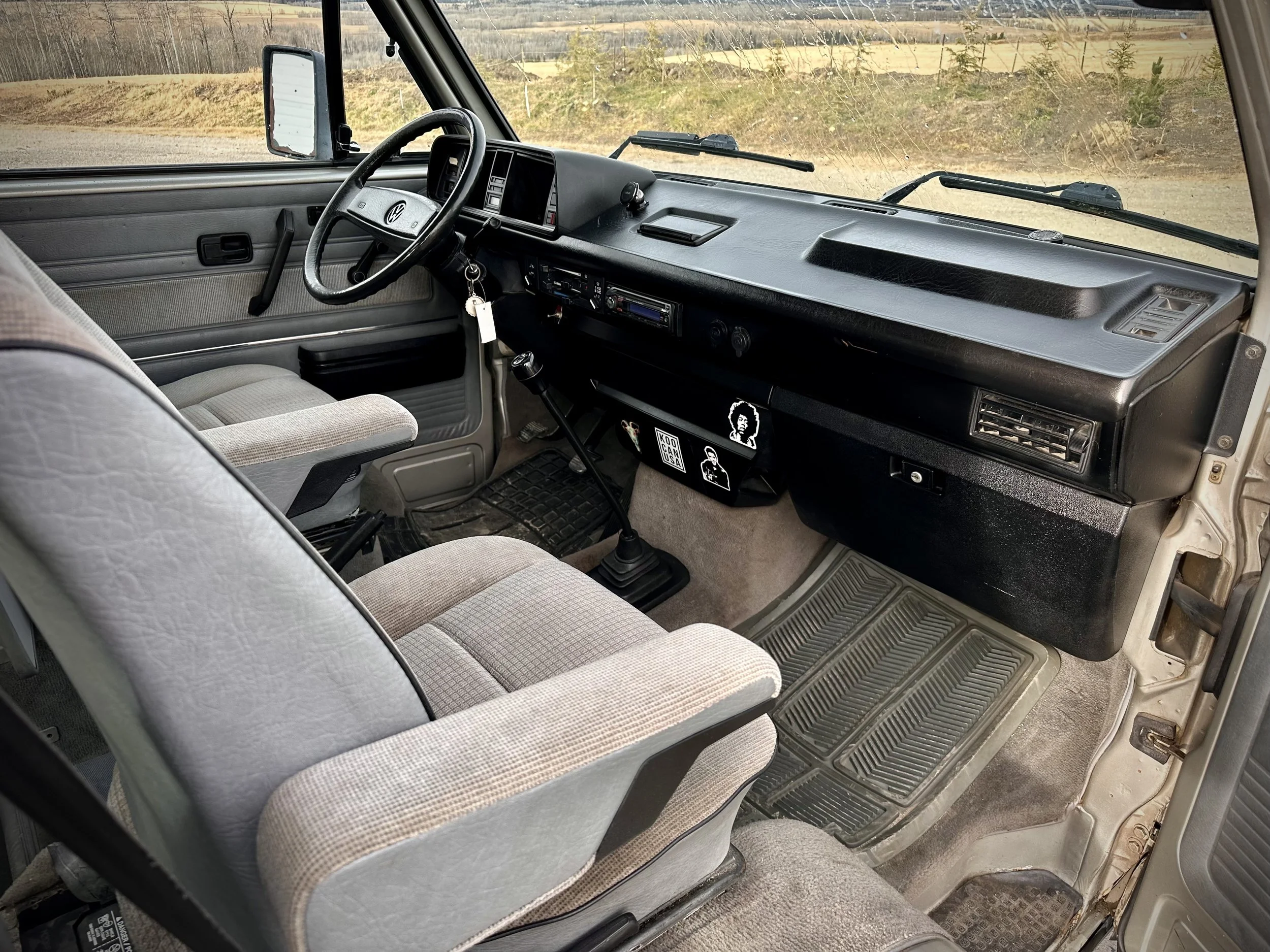 Inside the cab of a Vanagon camper van, showing the dashboard, steering wheel, gear shift, and two front seats with fabric upholstery, with a view of the outside landscape through the windshield.