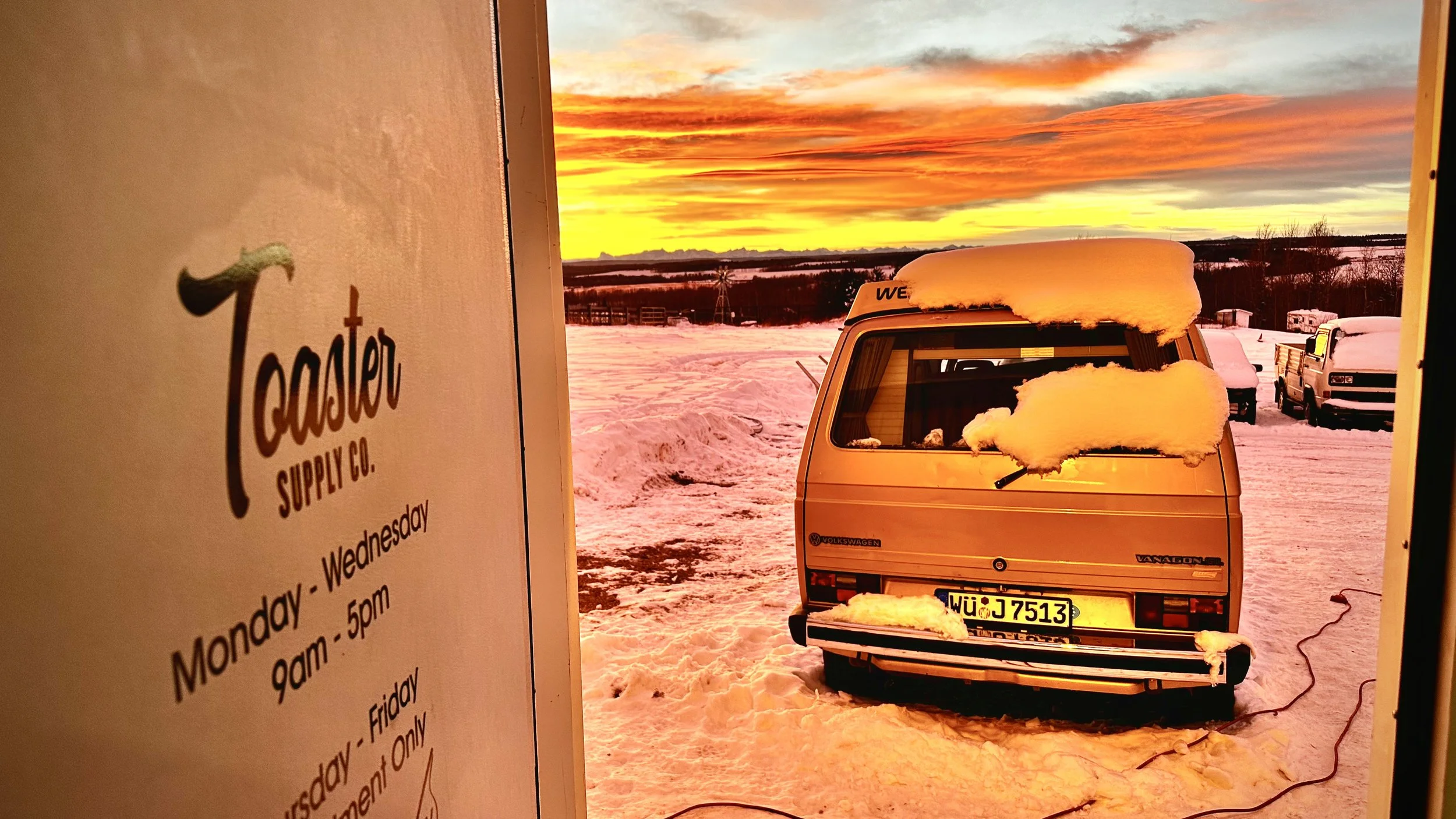 View through a doorway showing a snow-covered landscape with a vintage Volkswagen Vanagon vehicle and other parked cars, with a colorful sunset sky in the background.