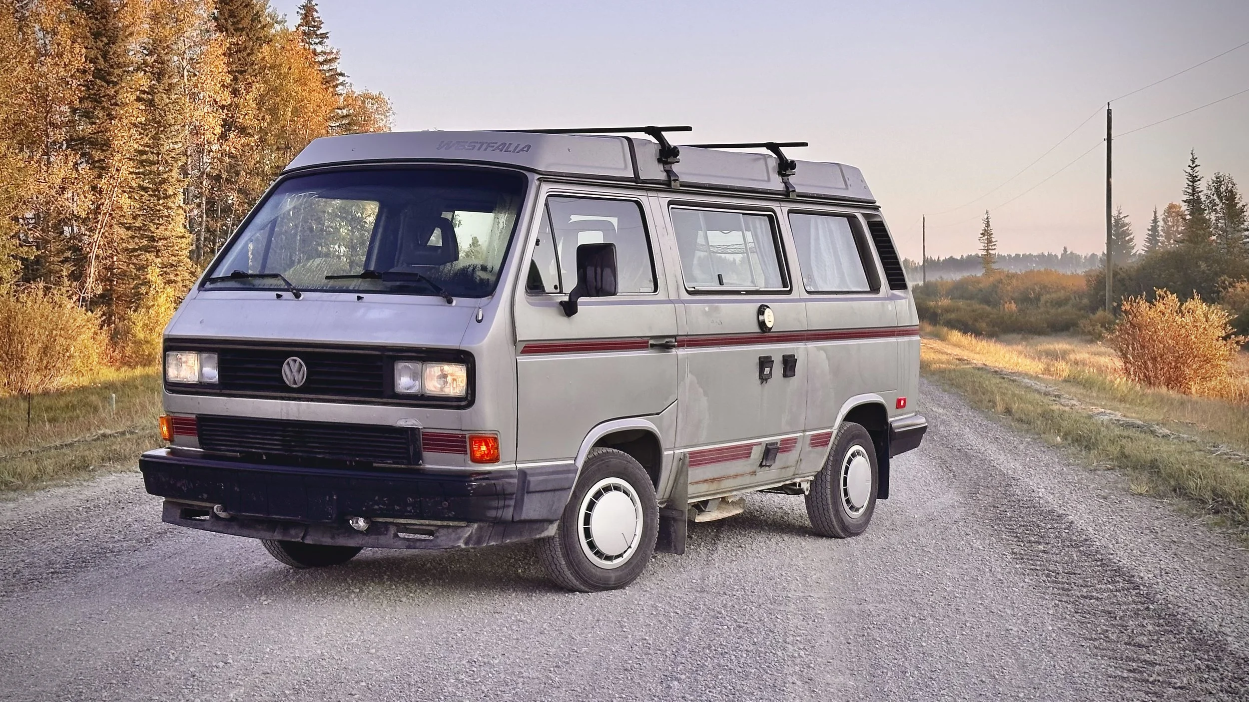 A vintage Volkswagen Vanagon camper van parked on a gravel road with autumn trees and a clear sky in the background.