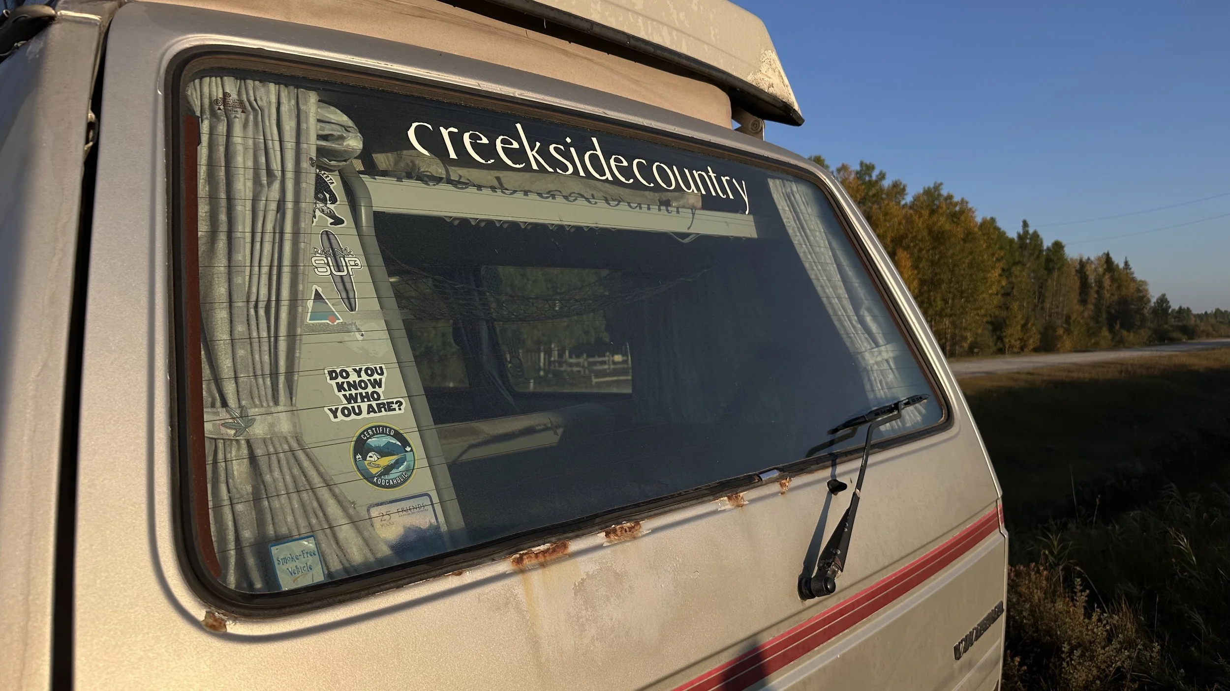 Close-up of a vintage Vanagon camper van with a large window, rust on the body, and various stickers. The van is parked next to a wooded area during sunset.