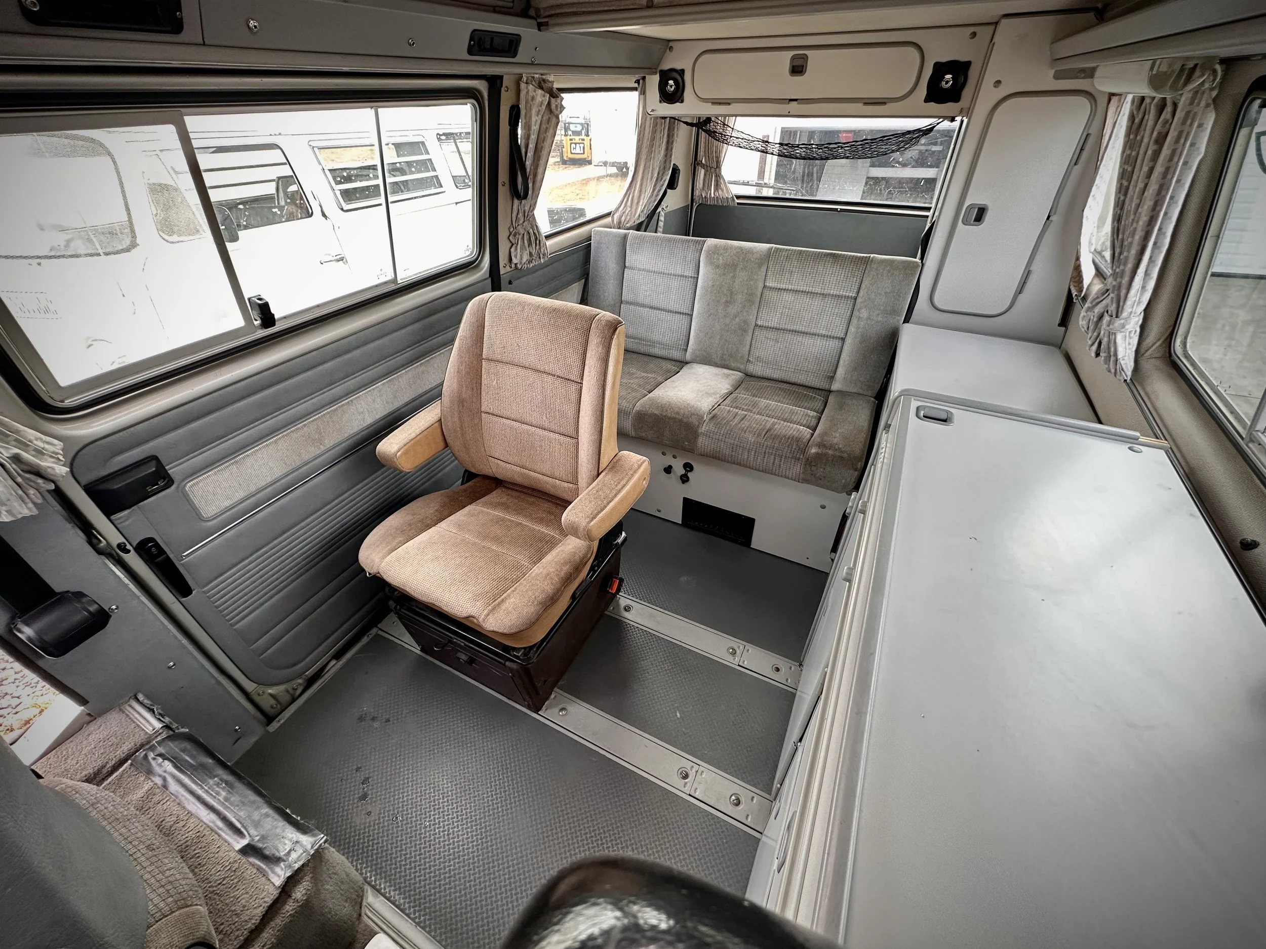 Interior of a Vanagon camper van with a tan driver's seat, a gray cushioned bench, a small white cabinet, and windows with curtains.