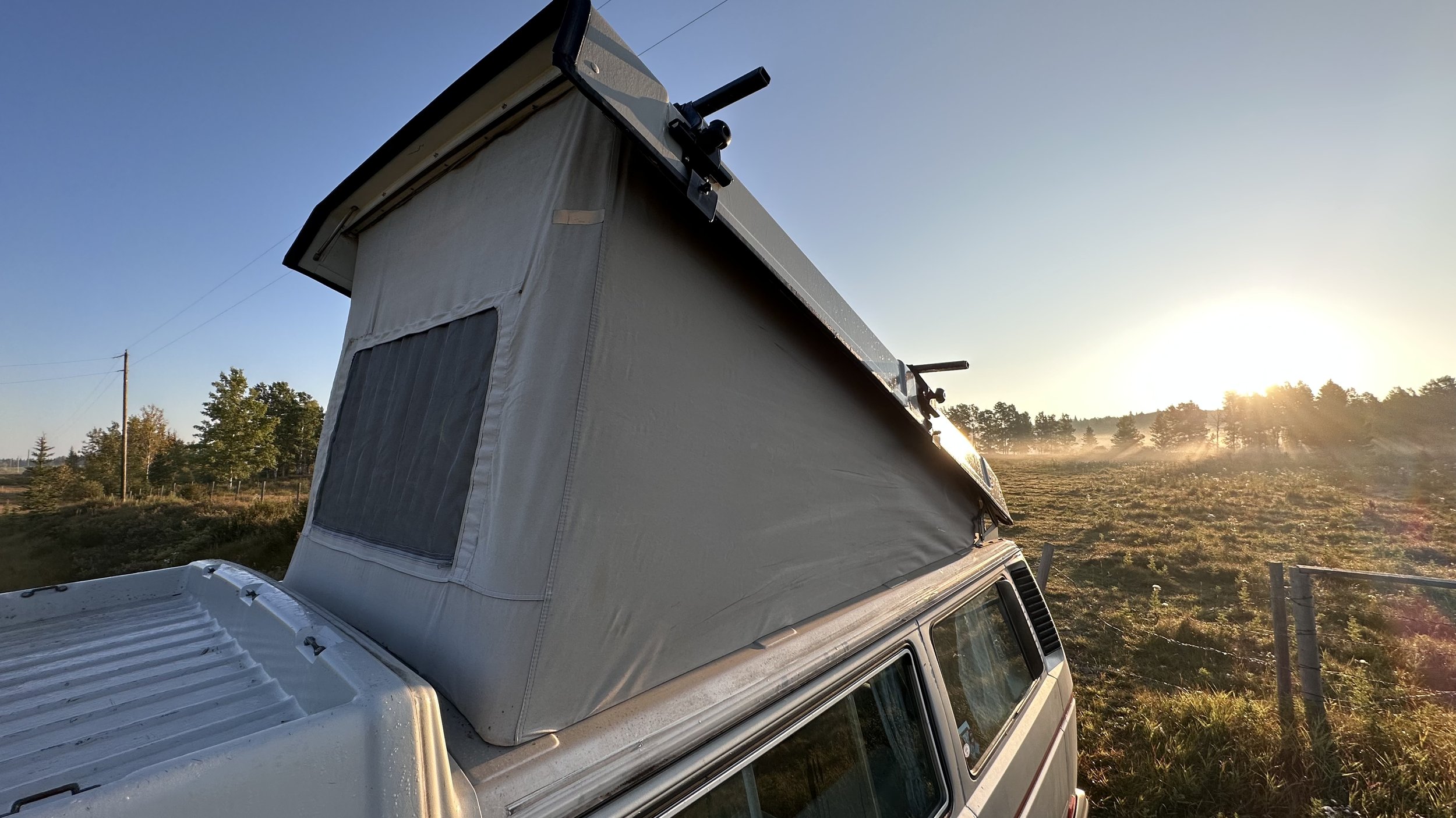 A Vanagon camper van with a pop-up roof tent is parked in a grassy area during sunset, with trees and a fence in the background.
