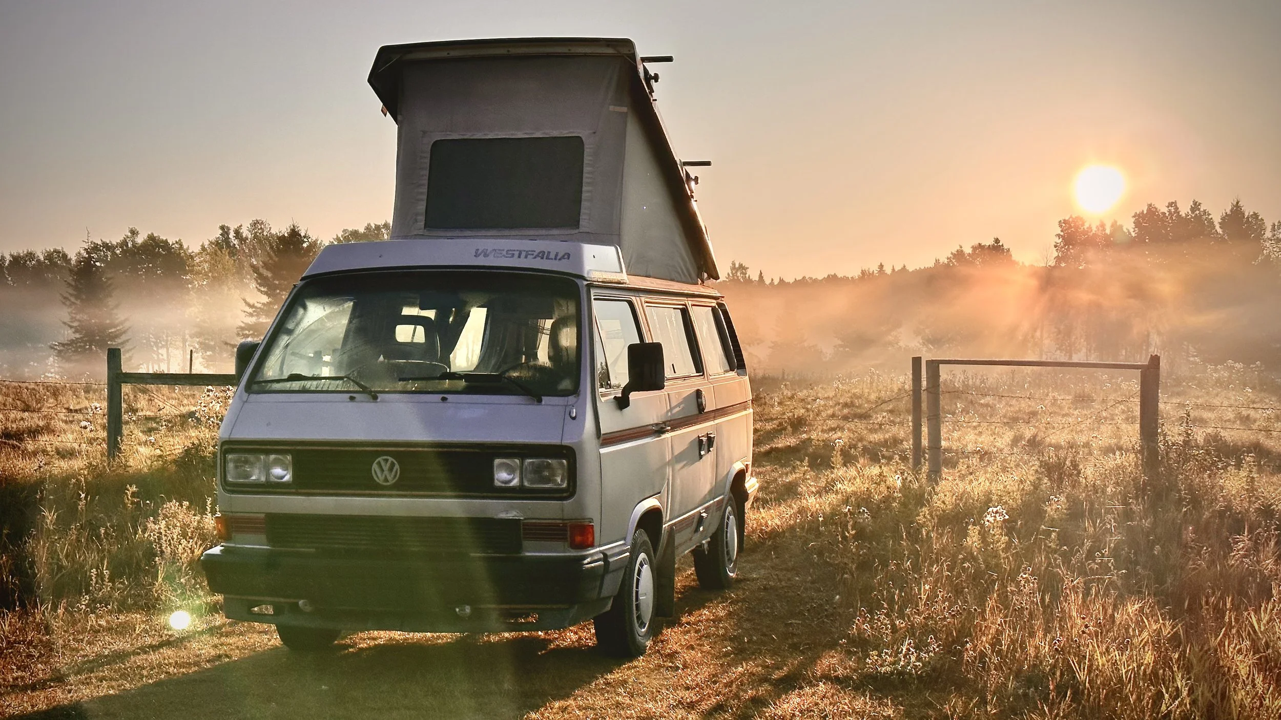 Vintage Volkswagen camper van with a pop-up roof parked on a grassy area during sunrise or sunset, with trees and a fence in the background.