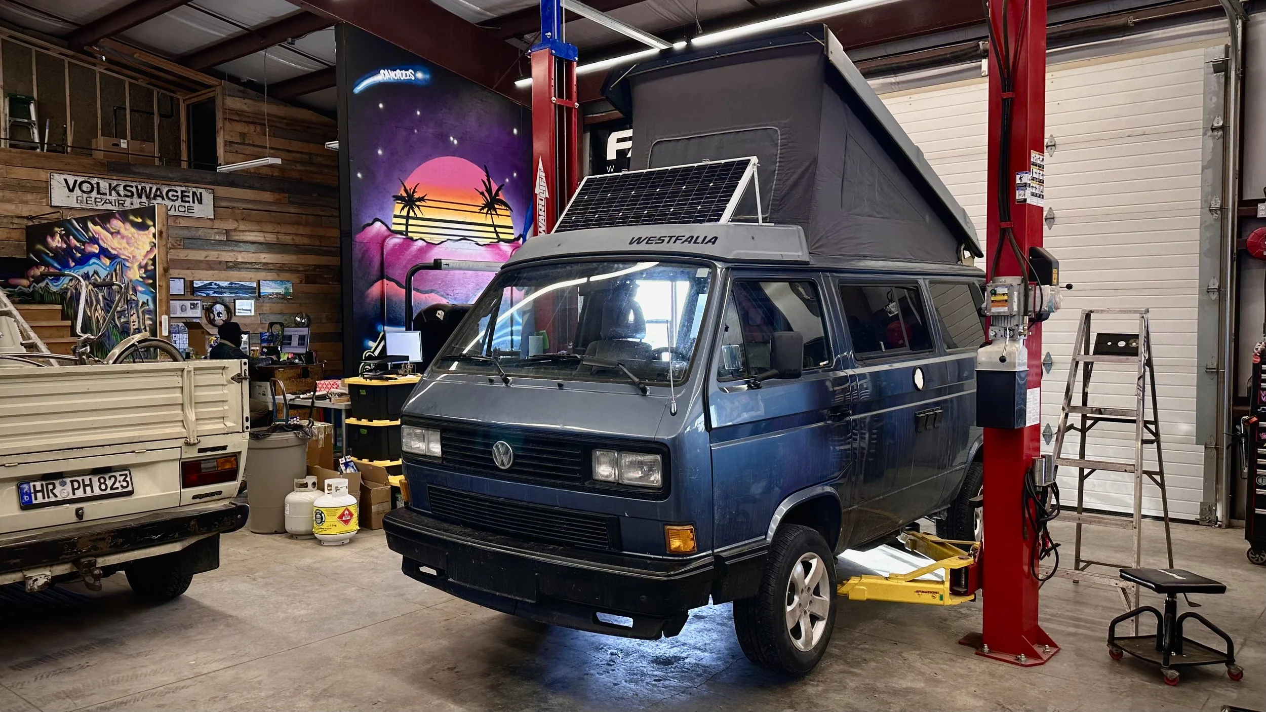 A blue Volkswagen Vanagon camper van on a hydraulic lift inside a garage, with a raised pop-up roof and solar panel on top.