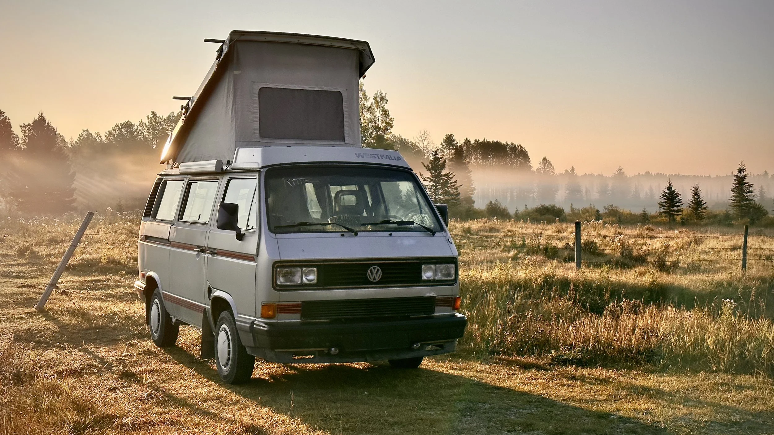 A vintage Volkswagen Vanagon with a pop-up roof parked on a grassy field during sunrise or sunset.
