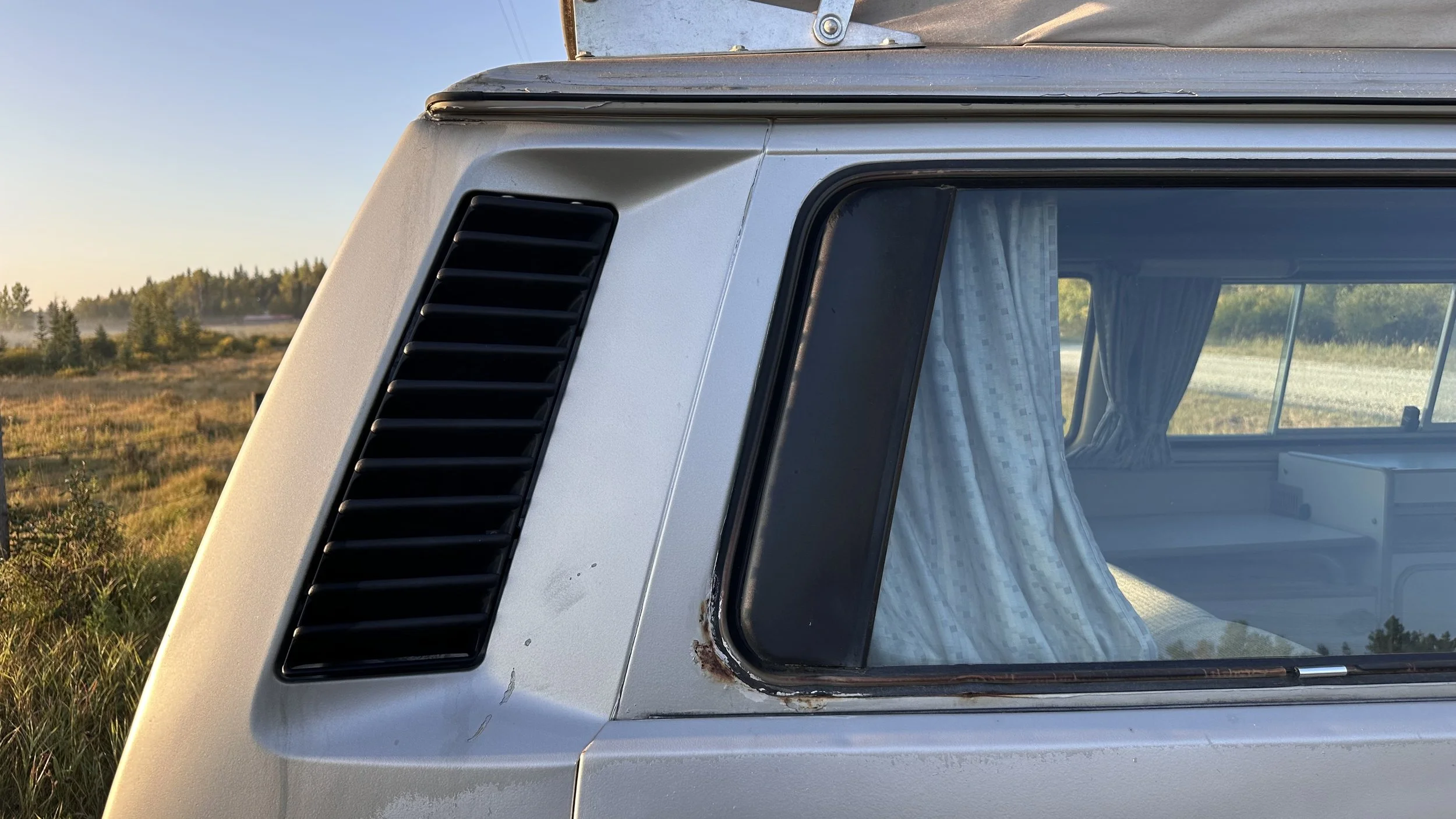 Close-up of the front corner of a vintage silver Vanagon camper van with a black vent and a window with curtains, parked in a grassy outdoor area.