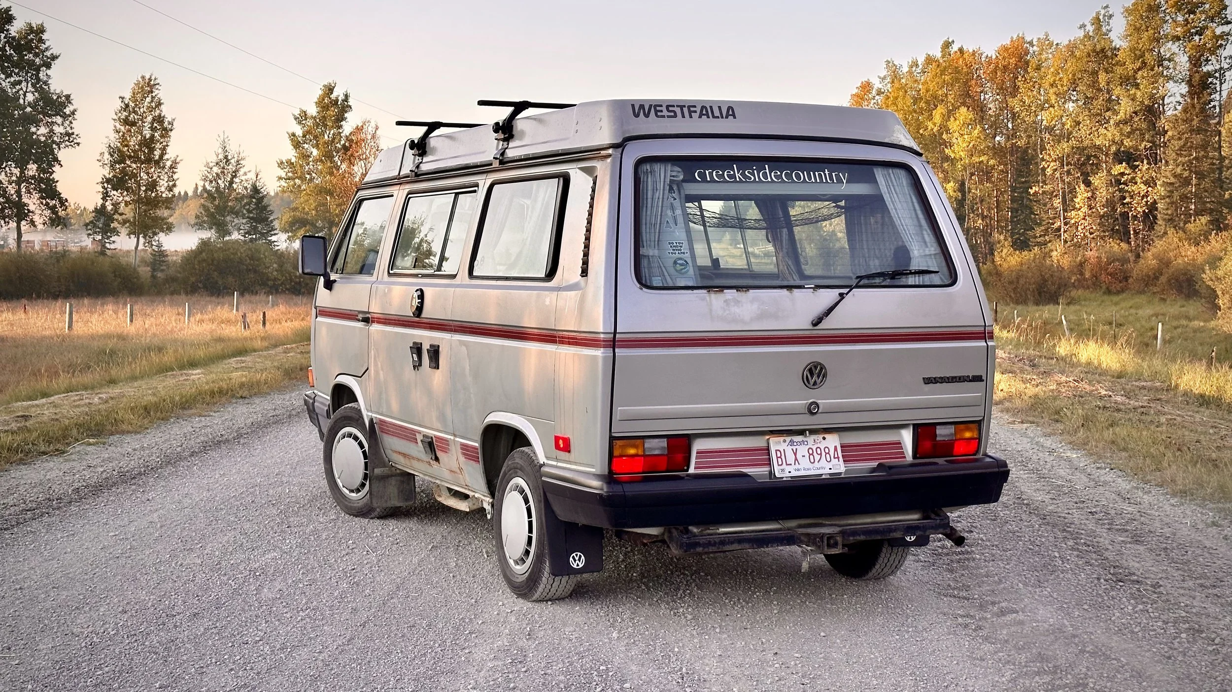 A vintage Volkswagen van parked on a gravel road with autumn trees in the background.