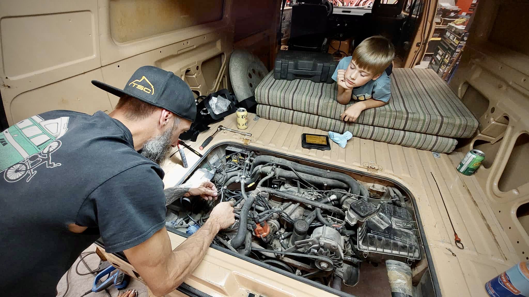 A man with a beard working on the engine of Vanagon with the hood open, while a young boy watches from a mattress placed inside the vehicle's interior.