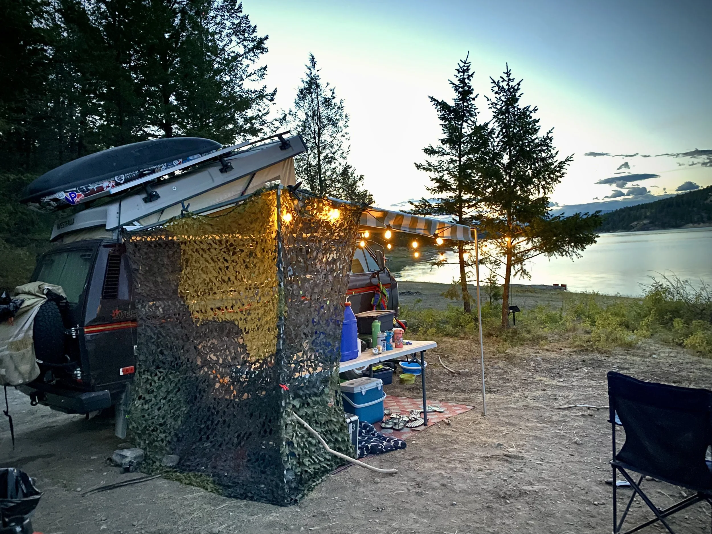 A camping setup by a lake during evening with a van, a table with supplies, string lights, and a camouflage privacy screen, surrounded by trees.