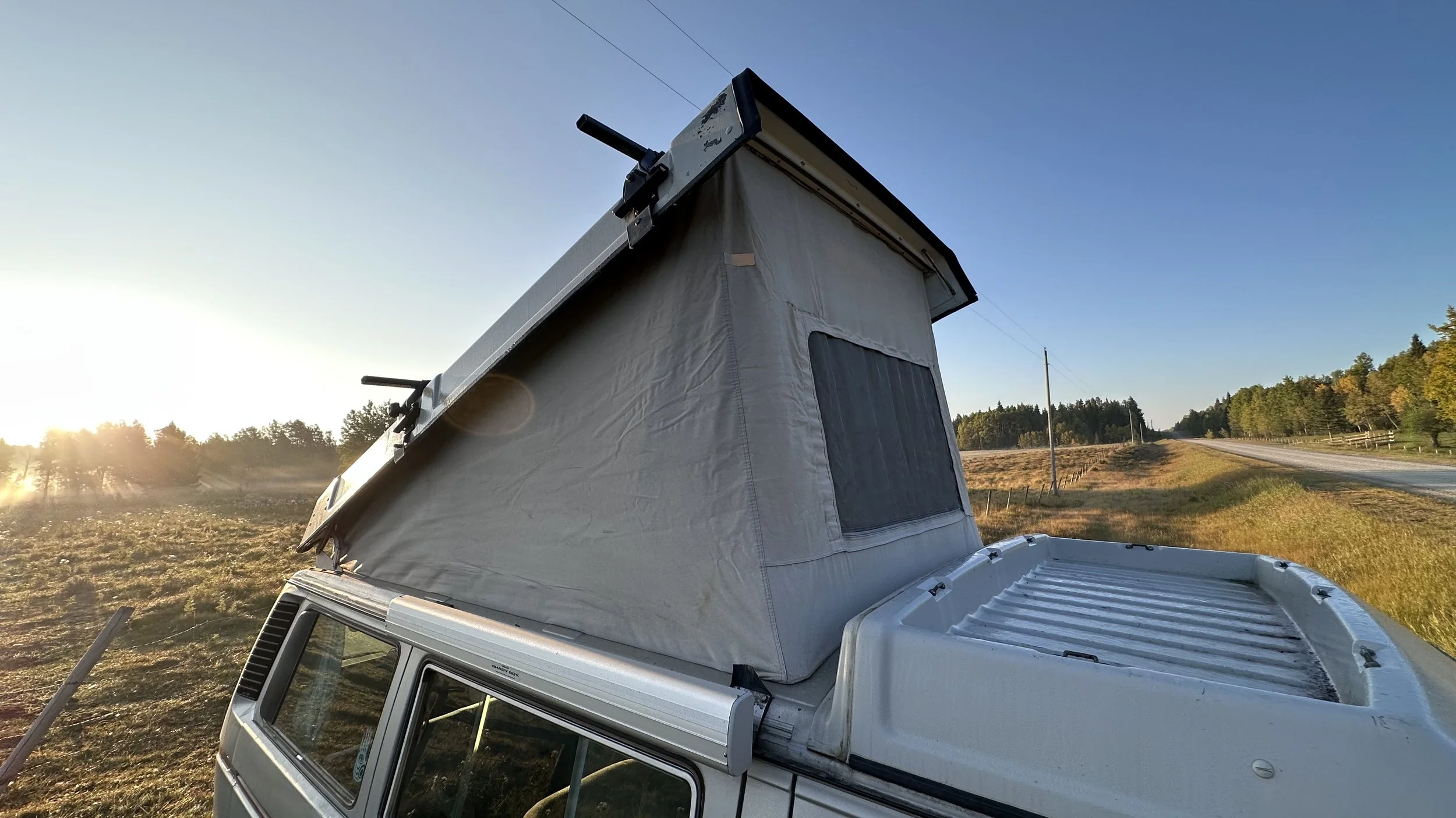 A silver Vanagon camper van with a raised pop-up roof tent on a rural road during sunset, surrounded by grassy fields and trees in the background.
