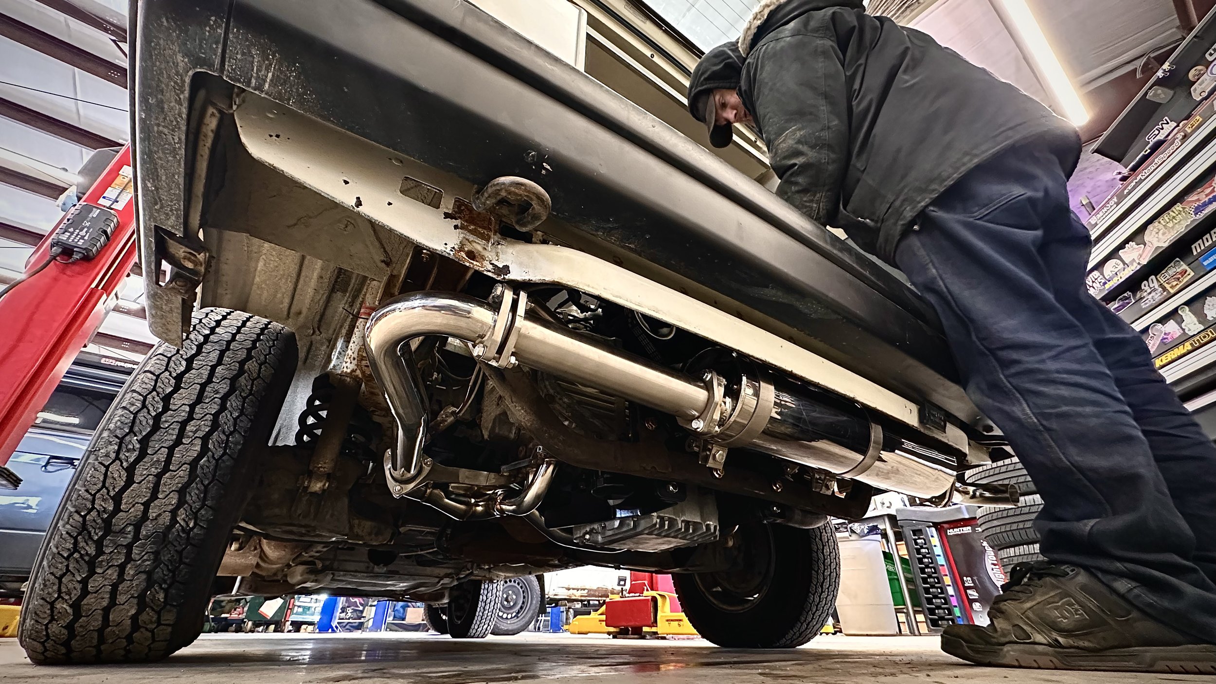 A mechanic working on the engine of a Vanagon in an auto workshop. The underside of the vehicle shows chrome exhaust pipes and suspension components. The mechanic is leaning forward, focused on his work.