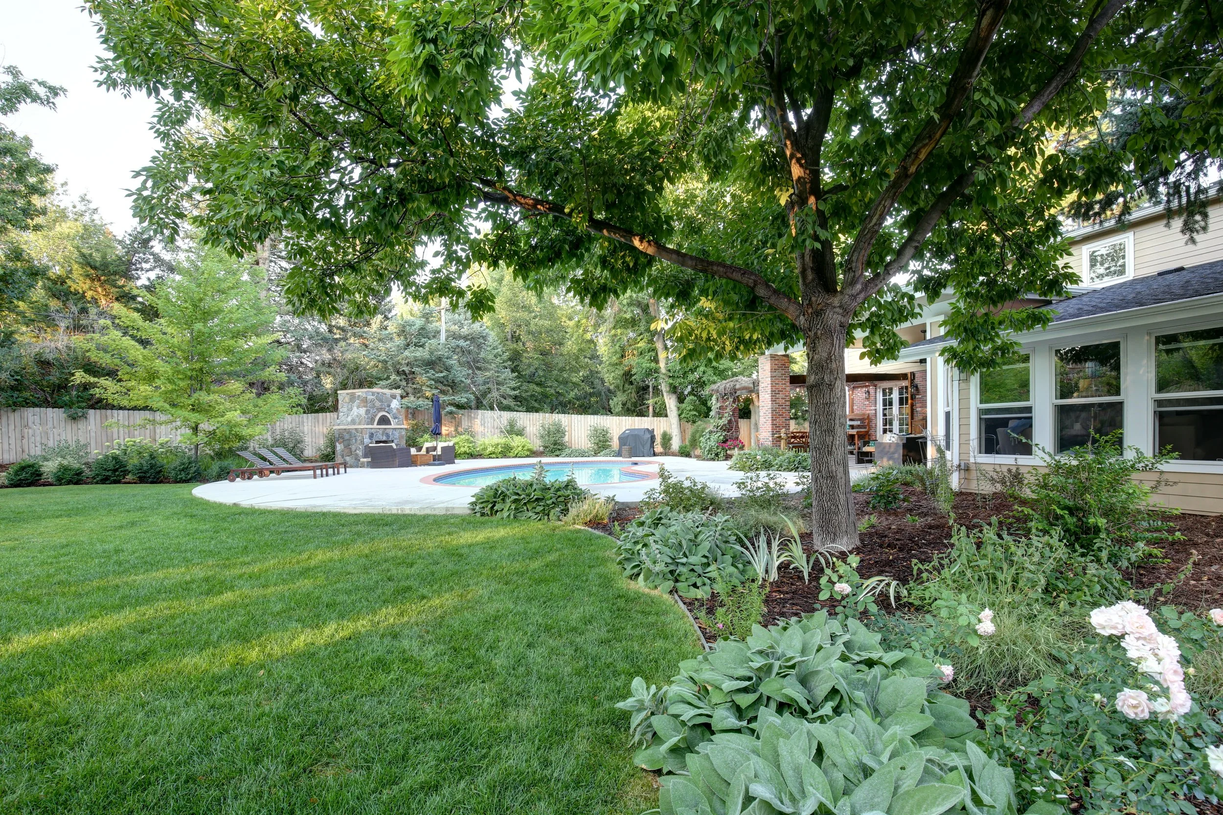 Denver backyard designed by The Green Fuse featuring a lush lawn, shade tree, pool, and stone outdoor fireplace with seating.
