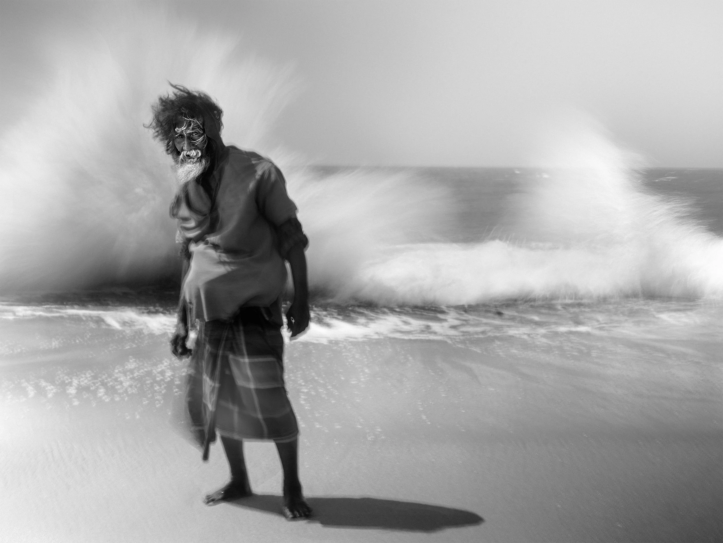 Black and white photo of a person standing on a beach with waves crashing in the background, wearing traditional clothing.