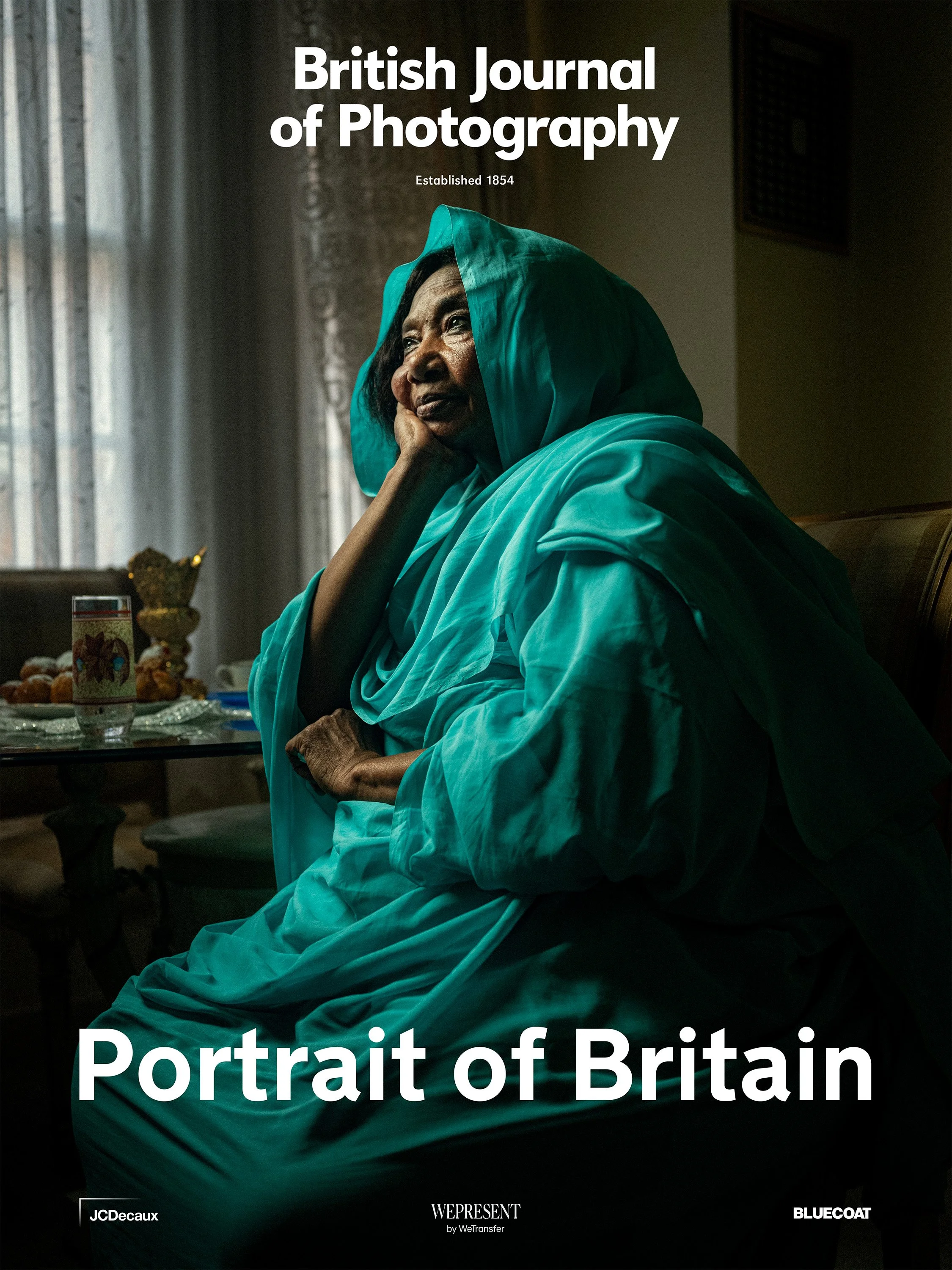 A portrait of Liela Medani seated in her London home, wearing a turquoise headscarf, resting her head on her hand and gazing thoughtfully toward a window.