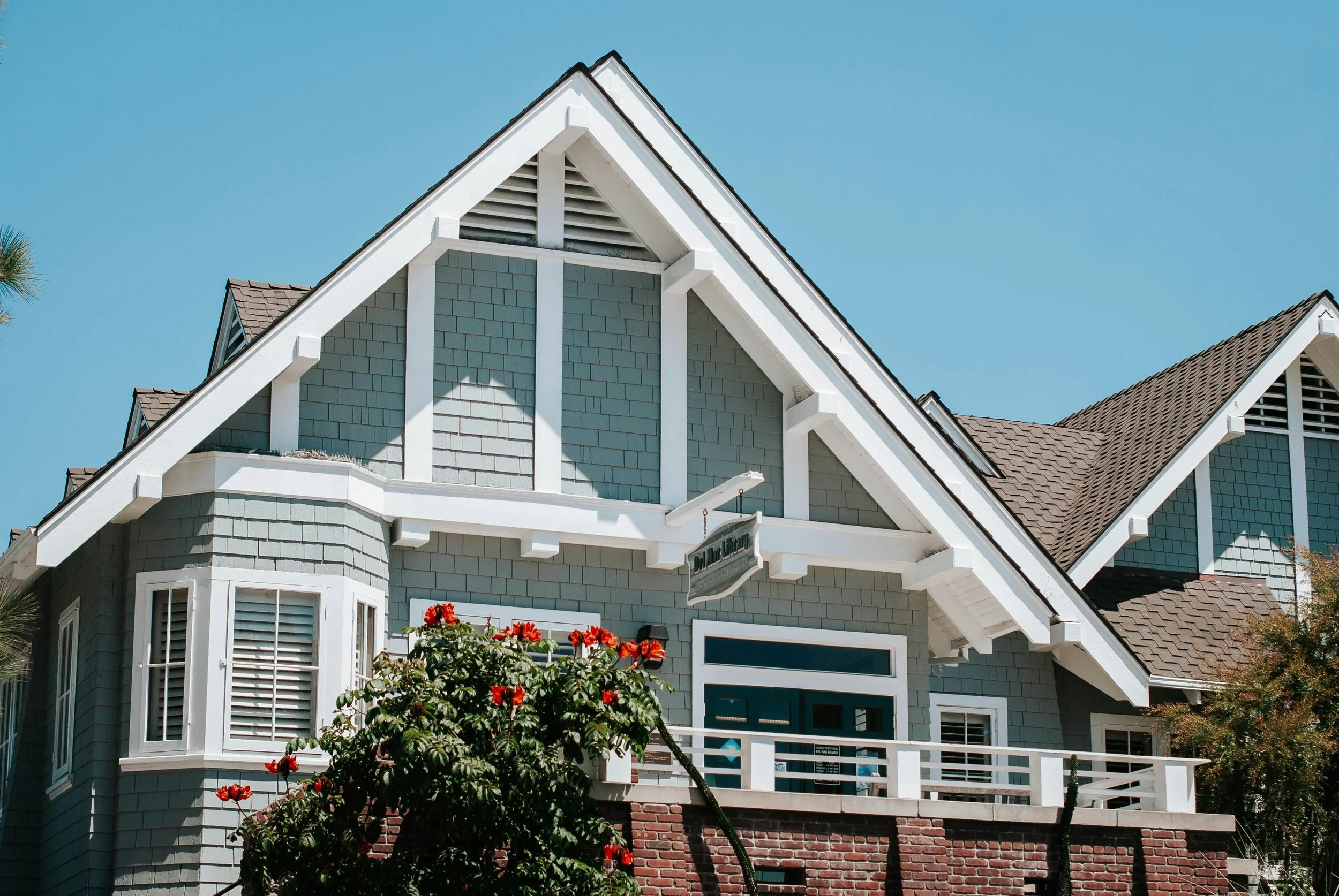 A two-story house with blue-green shingles, white trim, and a gabled roof with brown shingles. There is a balcony with white railing, a large bay window with shutters, and a sign hanging outside the house. A tree with red flowers is in front of the house.