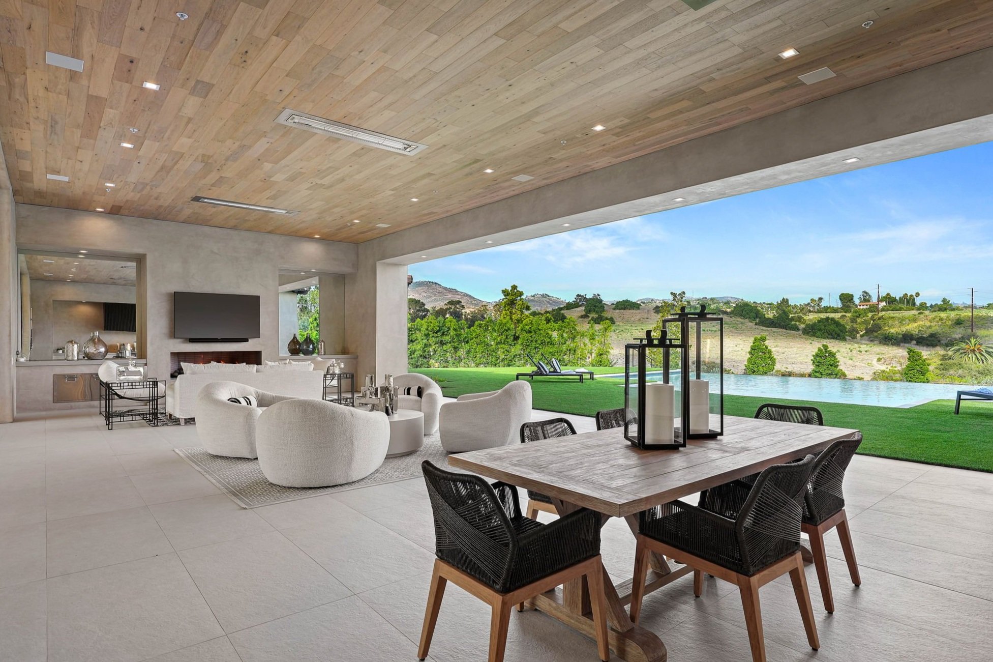 Modern living space with large open sliding doors revealing a lush green landscape with trees and hills, featuring a wooden ceiling, neutral-colored walls and flooring, and contemporary furniture including a dining table with black chairs and a cozy seating area with white armchairs and a TV.