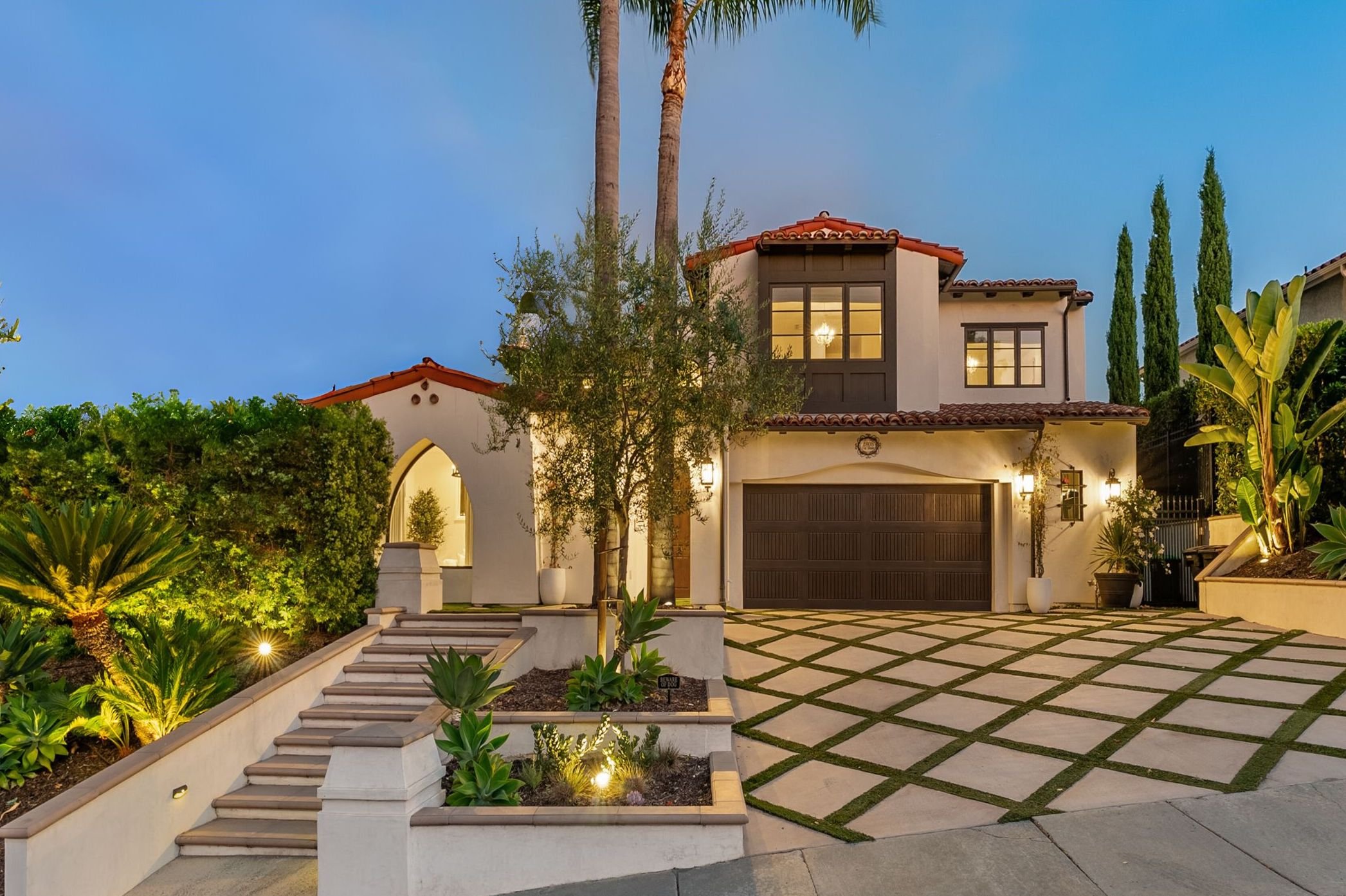 Luxury two-story house with a red-tile roof, dark brown garage door, and night lighting, surrounded by lush landscaping with tall trees and plants.