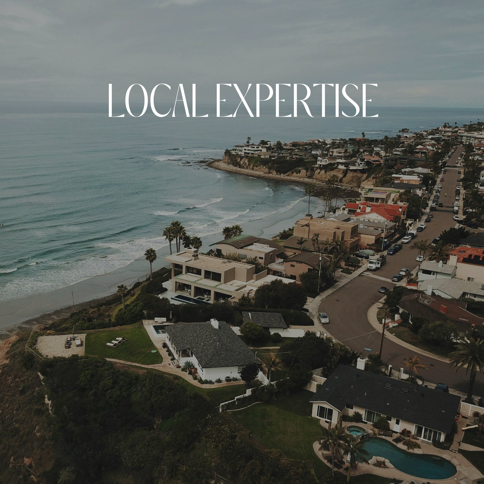 Aerial view of a coastal neighborhood with beachfront houses, palm trees, and a curving road along the shoreline, under a cloudy sky.