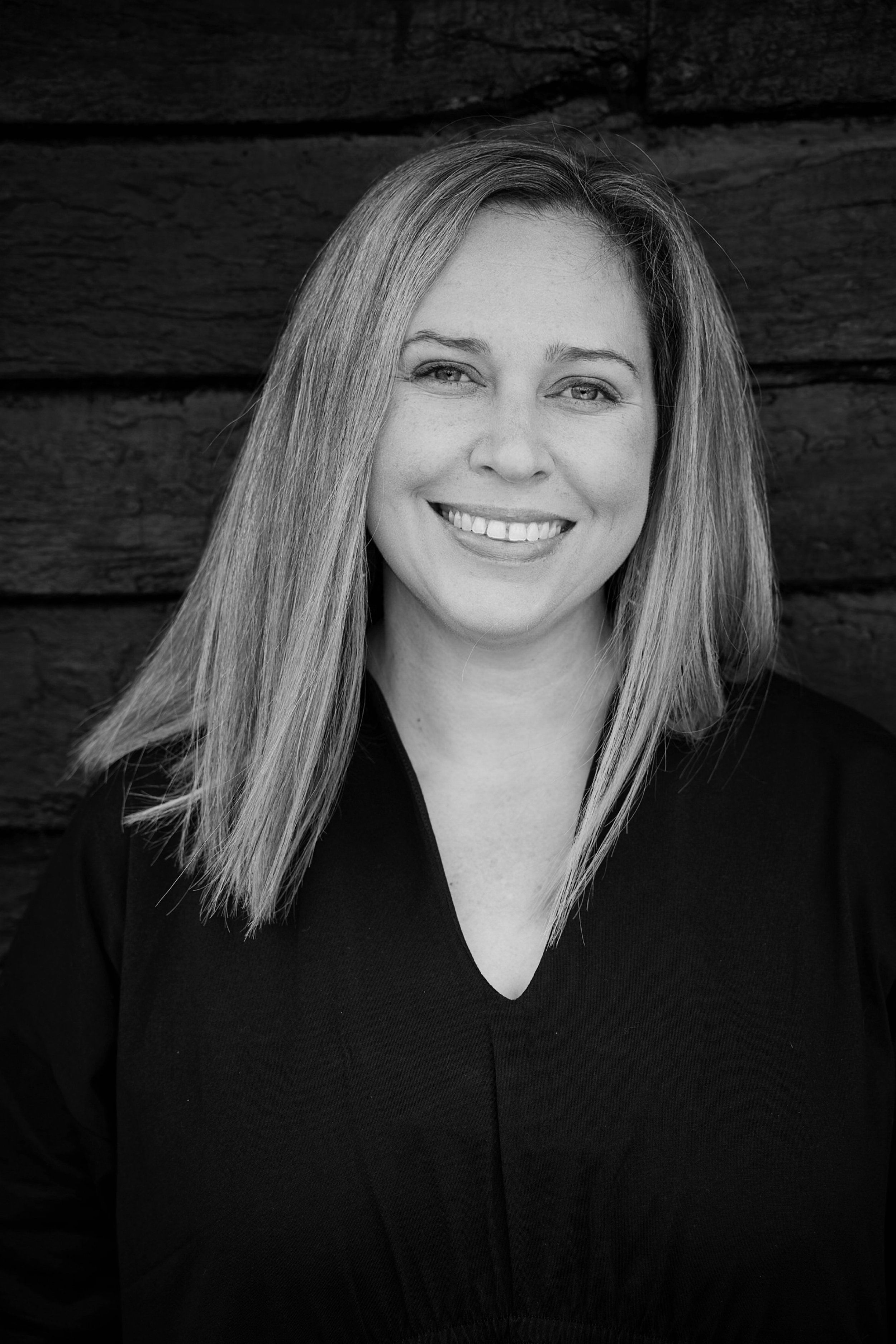 Black and white portrait of a smiling woman with long straight hair, wearing a V-neck top, standing in front of a dark wooden background.