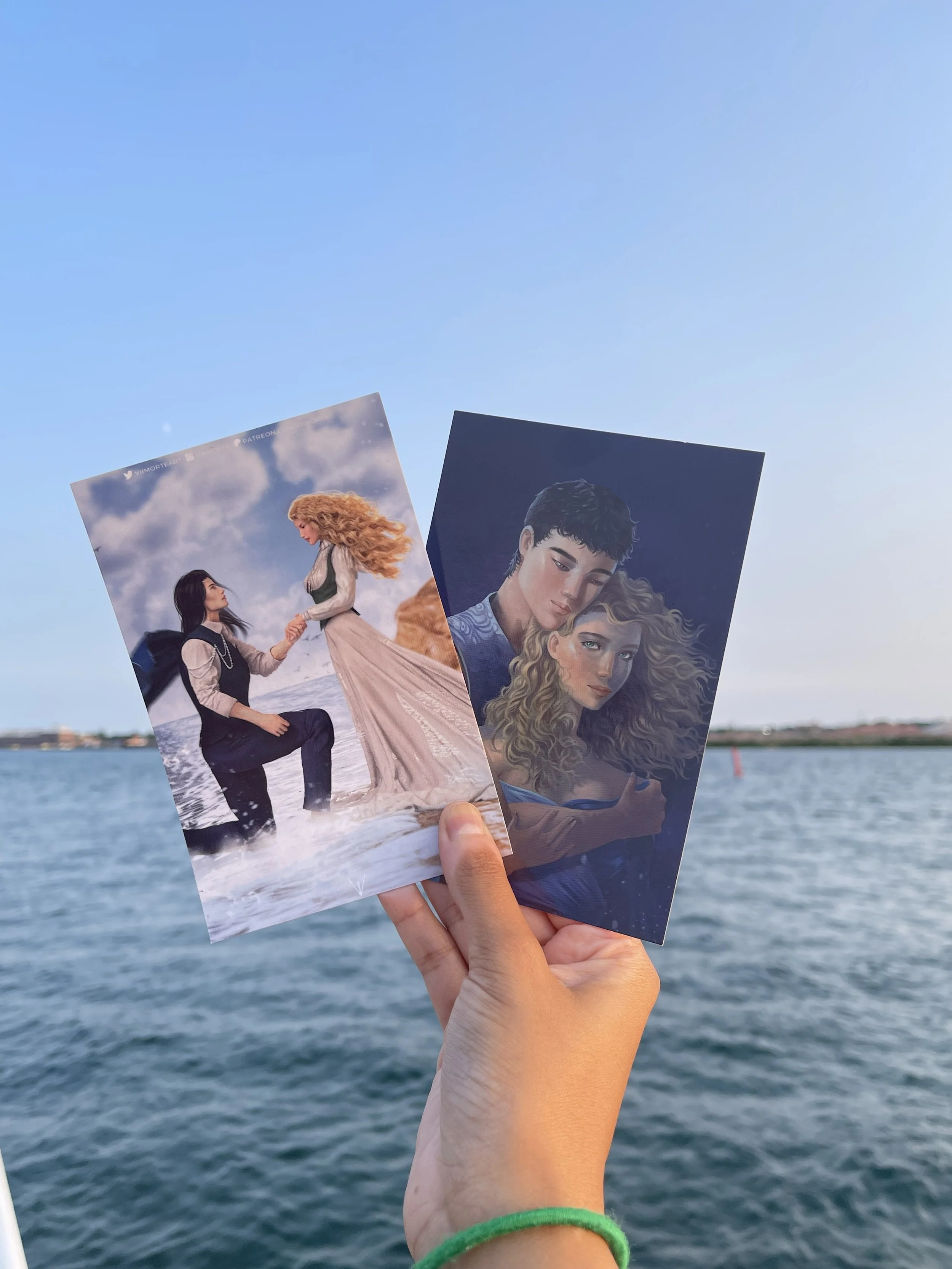 A hand holding two illustrated posters of women with a body of water and distant shoreline in the background.