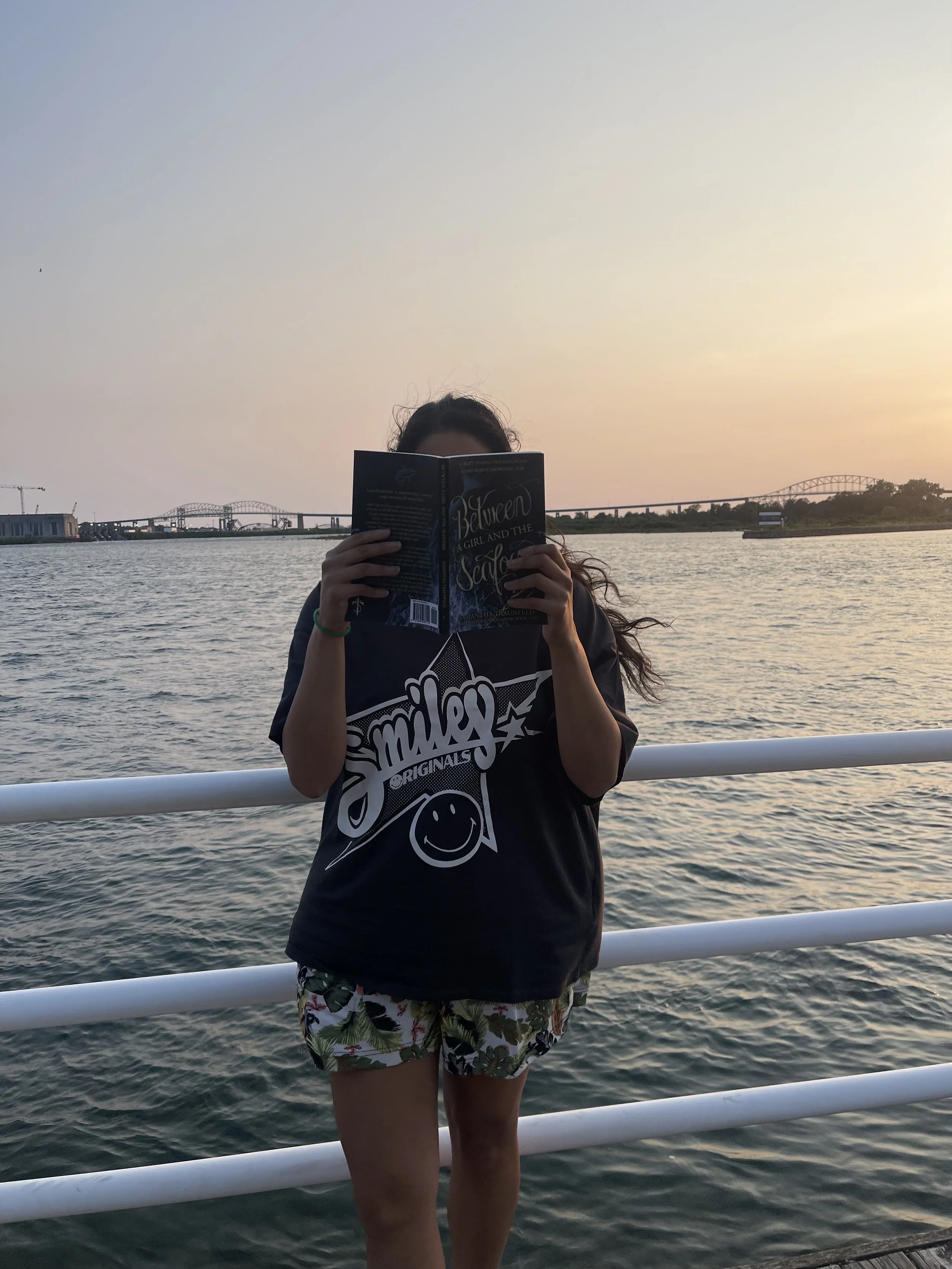Person with long hair wearing a black t-shirt with white text and colorful shorts, standing by water at sunset, reading a book.