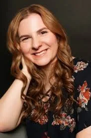 A woman with long, wavy, red hair smiling and posing with her hand resting on her face, wearing a dark floral top against a dark background.