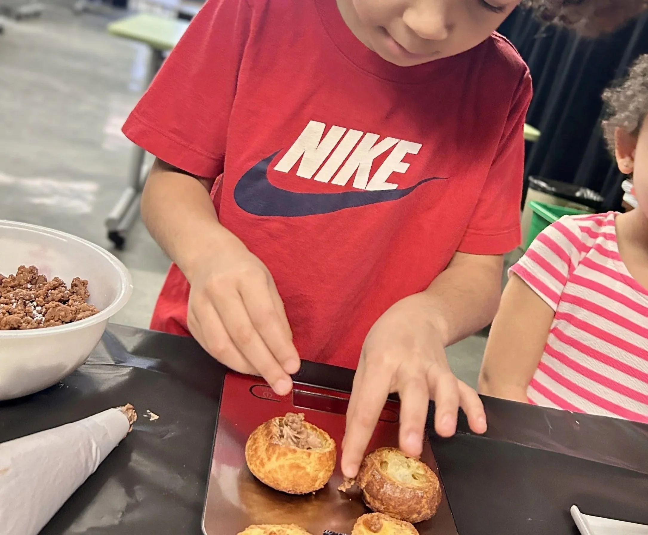 Un garçon avec des cheveux bouclés portant un t-shirt rouge Nike en train de préparer ou de manger des beignets dans une cuisine ou une salle de classe.