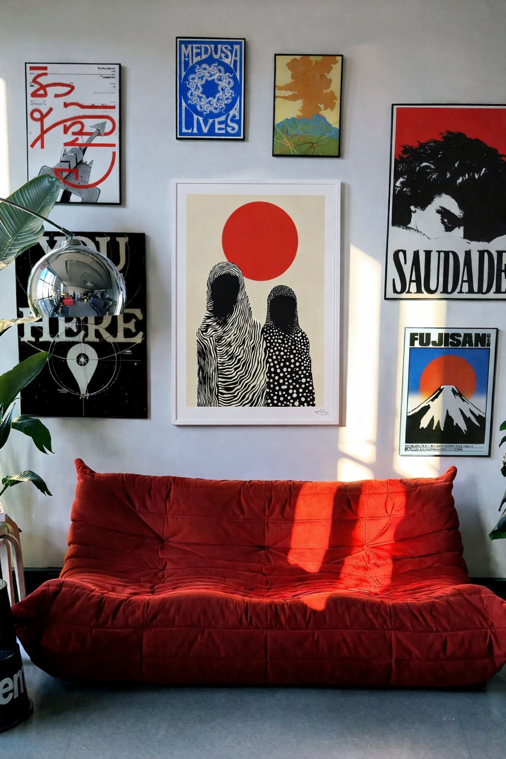 A red velvet couch in front of a white wall decorated with various framed art prints, including images of Mount Fuji, a red sun, and stylized families, with plants and a metallic globe-like lamp nearby.
