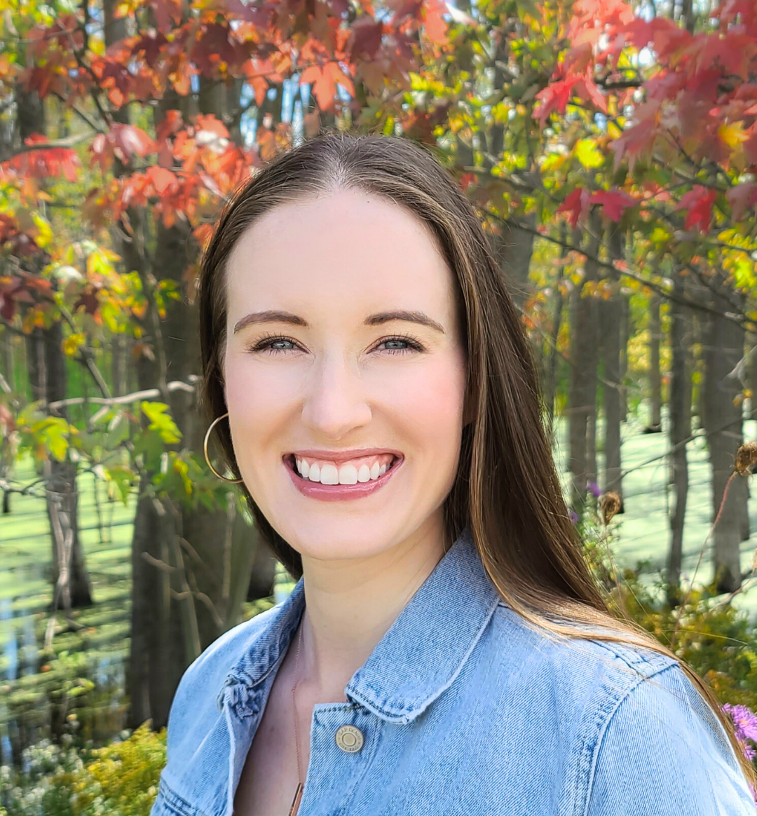 A woman smiling outdoors in front of trees with red and green leaves, wearing a denim jacket and hoop earrings.