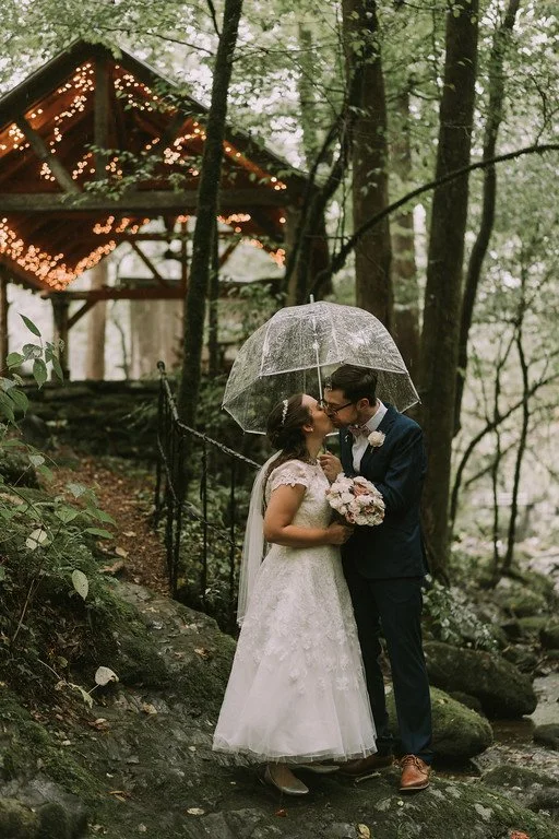 The couple stands down the hill from the wedding pavilion, which is lit by warm string lights. They share a kiss under a clear umbrella.