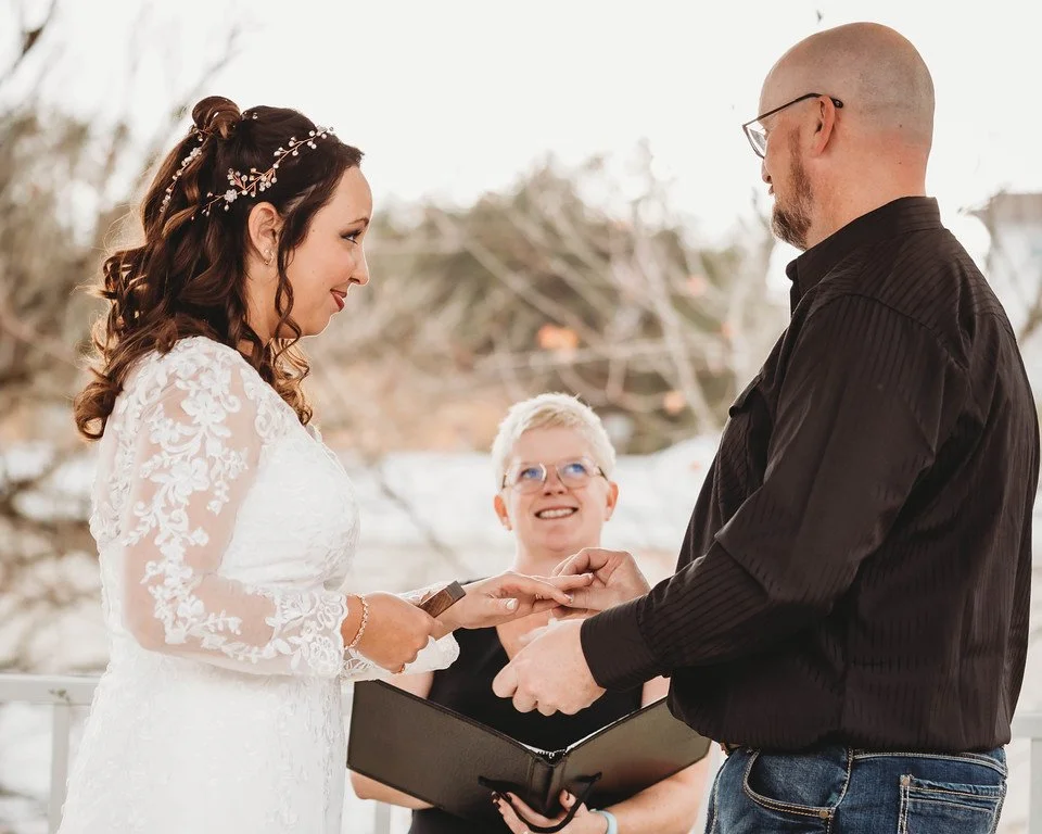 The couple stand facing eachother at the altar in front of the officiant. One puts a ring on the other. 