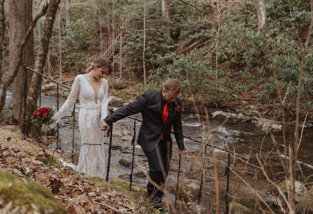 The couple walk, holding hands, down a tricky path beside a flowing river in the woods. They are almost facing the camera.