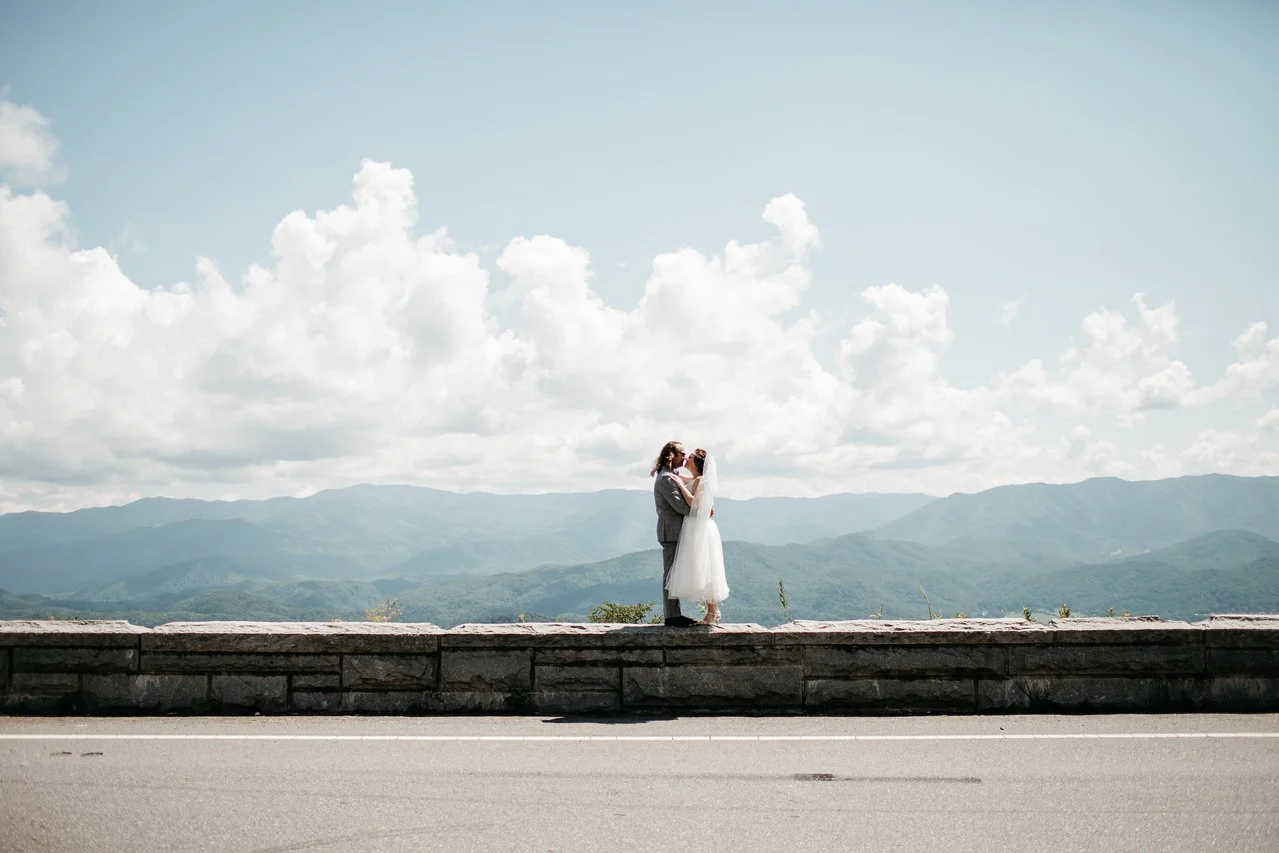 The couple shares a kiss standing on a short stone wall beside the road. Behind them are misty mountains, fluffy white clouds, and a bright blue sky.