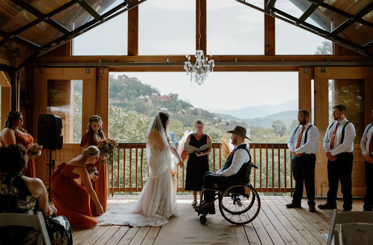 The bride stands facing the groom who sits in a wheelchair facing the bride. In between them the officiant is seen reading from her folder. On either side of the couple are bridesmaids and best men. The background overlooks the mountains.