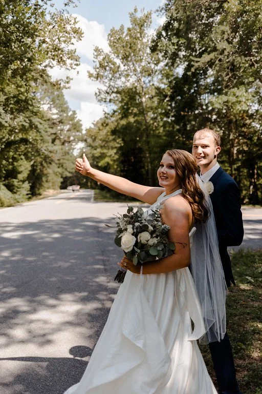 The couple stand at the side of the road in the shade. One puts their thumb out as if hitchhiking. They are both smiling, and in the background, the road leads into the dense green forest.