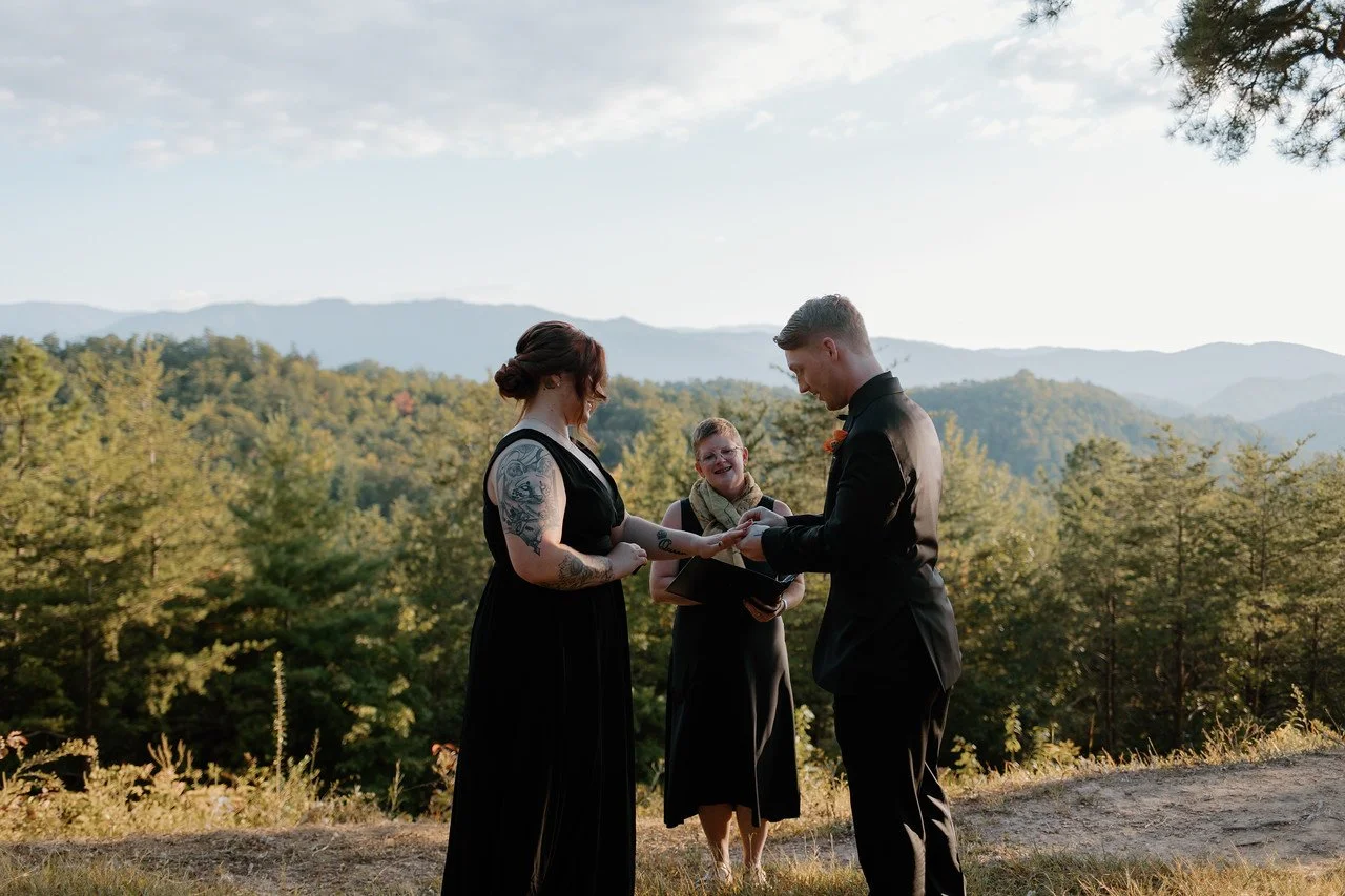 The couple stand facing eachother in front of the officiant, while one puts a ring on the other. Behind everyone is a view of the green and blue mountains in the distance, while the sun is moderately low in the sky just out of shot.