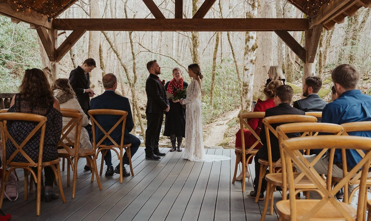 The couple stands facing eachother in front of the officiant at the altar in a pavilion. In the foreground are the wedding guests sitting on either side of the aisle. The pavilion overlooks a nearly bare forest and stream.
