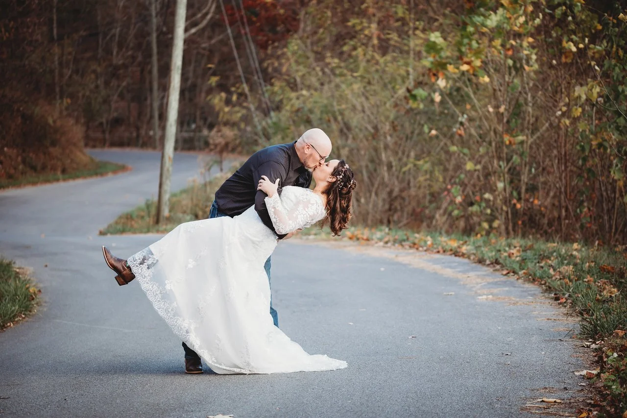 The couple perform a leaning kiss in the road. In the background is a telephone pole, and lots of vegetation.