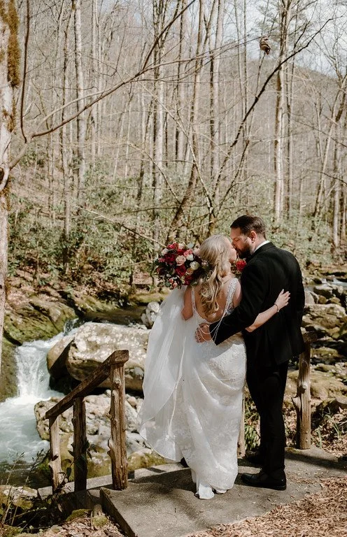 The couple stand at the top of a staircase, sharing a kiss with their backs to the camera. In front of them is a rushing stream and large rocks. Beyond that is a nearly bare forest.