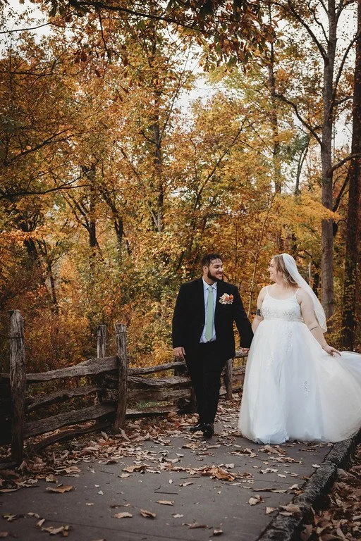 The couple walk down a leafy path in autumn. Around them are yellow leaves, and to the side of the path is a wooden fence. The couple look at eachother lovingly.