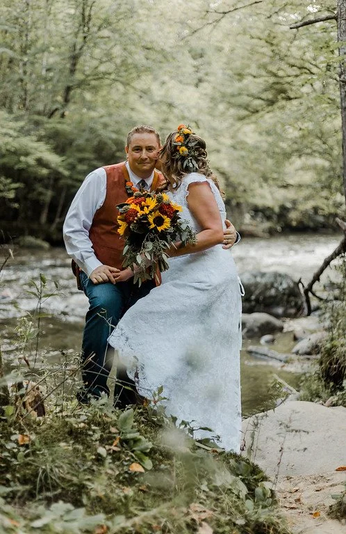 The couple stands, embracing, on large river rocks beside a flowing river. One kisses the other on the cheek while the other looks at the camera.