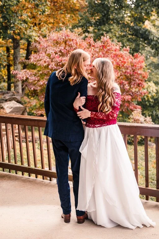 The couple stands, embracing, on the porch of a cabin, overlooking a yard with a bright pink tree. One faces the camera, while the other has their back to the camera, and they share a kiss.