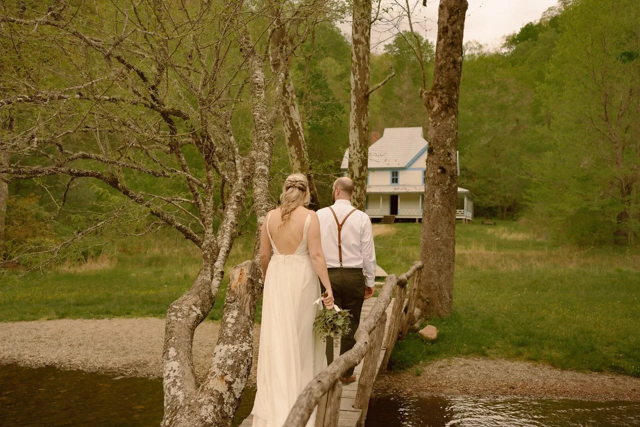 The couple walk across a thin wooden bridge across a small stream to a clearing the woods. In the clearing is an old white house with blue accents.