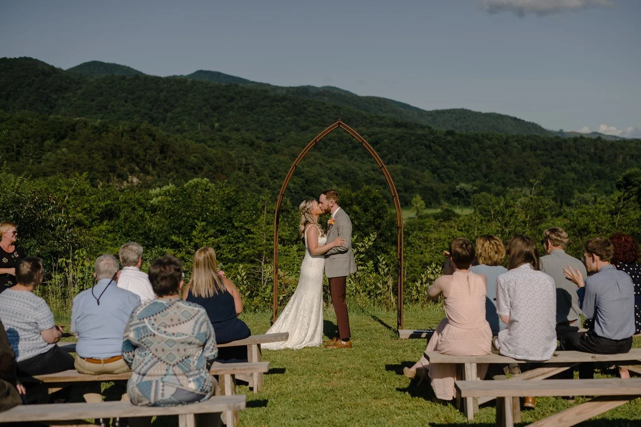 The couple stand under the wedding arch, sharing a kiss. In the foreground, wedding guests sit on either side of the aisle. Behind the altar are lush green mountains, and a clear sky. The sun shines down on the couple from behind the camera.