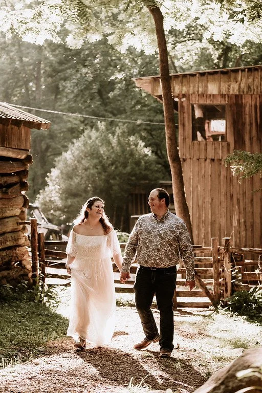The couple walk towards the camera holding hands. Behind them are various old wooden buildings and trees. The sun shines from behind them.