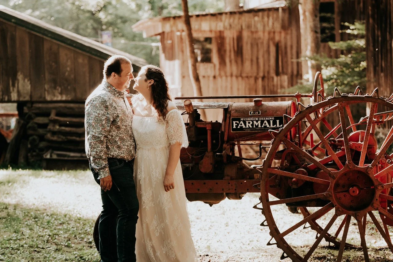 The couple stand embracing eachother in front of a red, antique piece of farming equipment. In the background are various old wooden buildings and many trees.