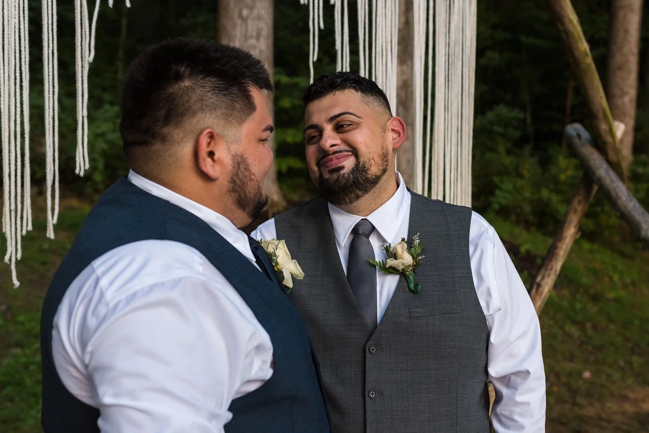 The couple look at eachother lovingly in front of the altar. Behind the altar are dark green leaves.